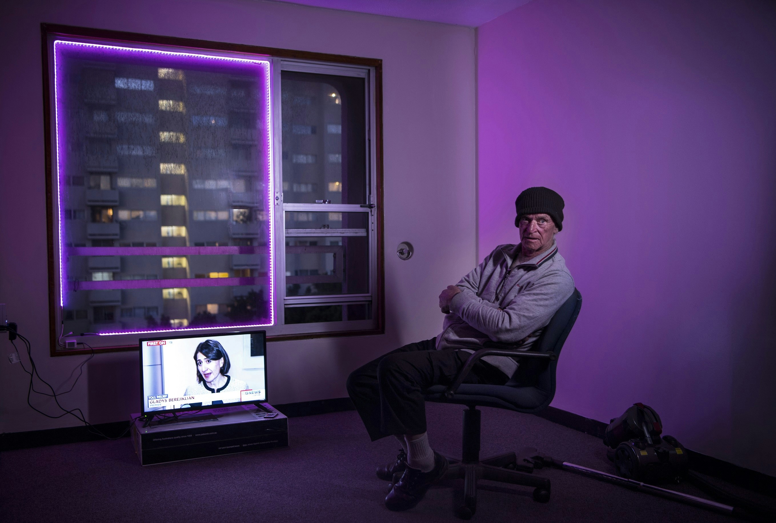 An elderly Caucasian man wearing a beanie and a grey sweater sits in a sparsely furnished room with purple fairylights fixed to the window frame. A television screen shows politician Gladys Berejiklian speaking at a press conference.
