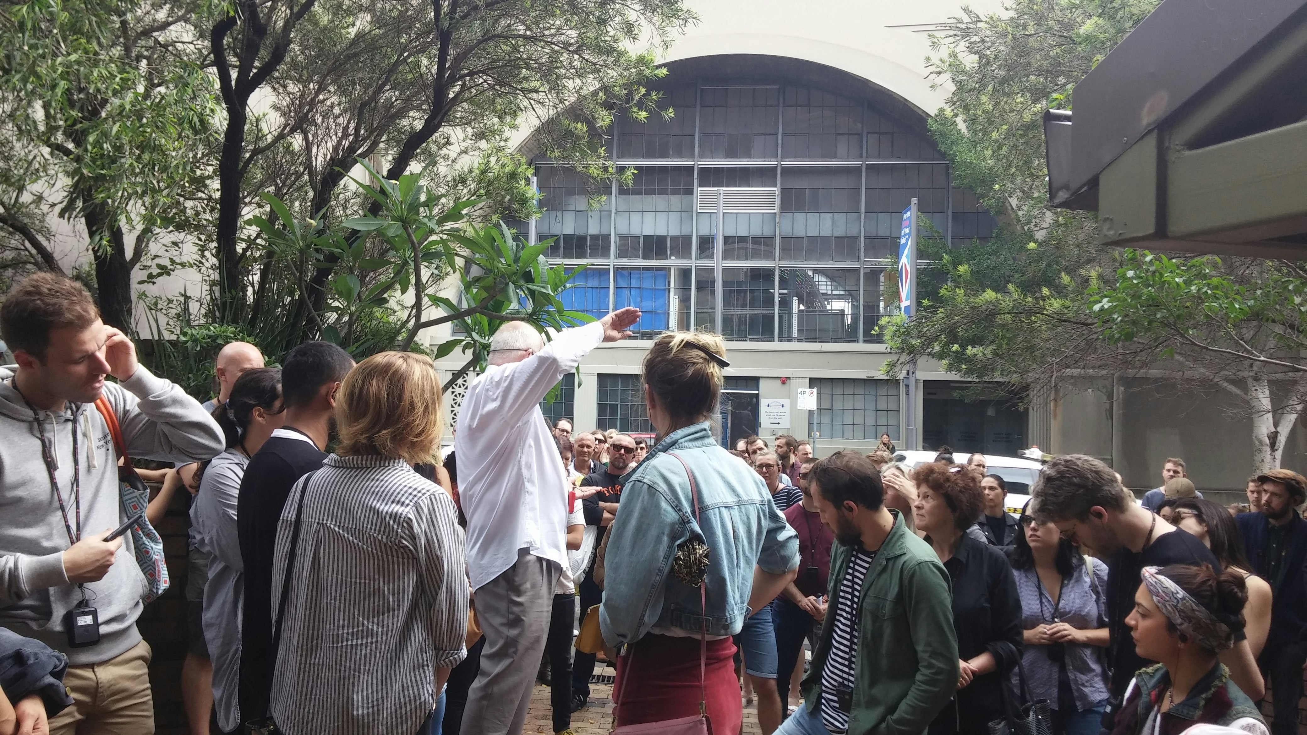 A crowd standing in an outdoor urban space, listening to an elderly male-presenting figure in a white dress shirt and grey trousers present a talk.