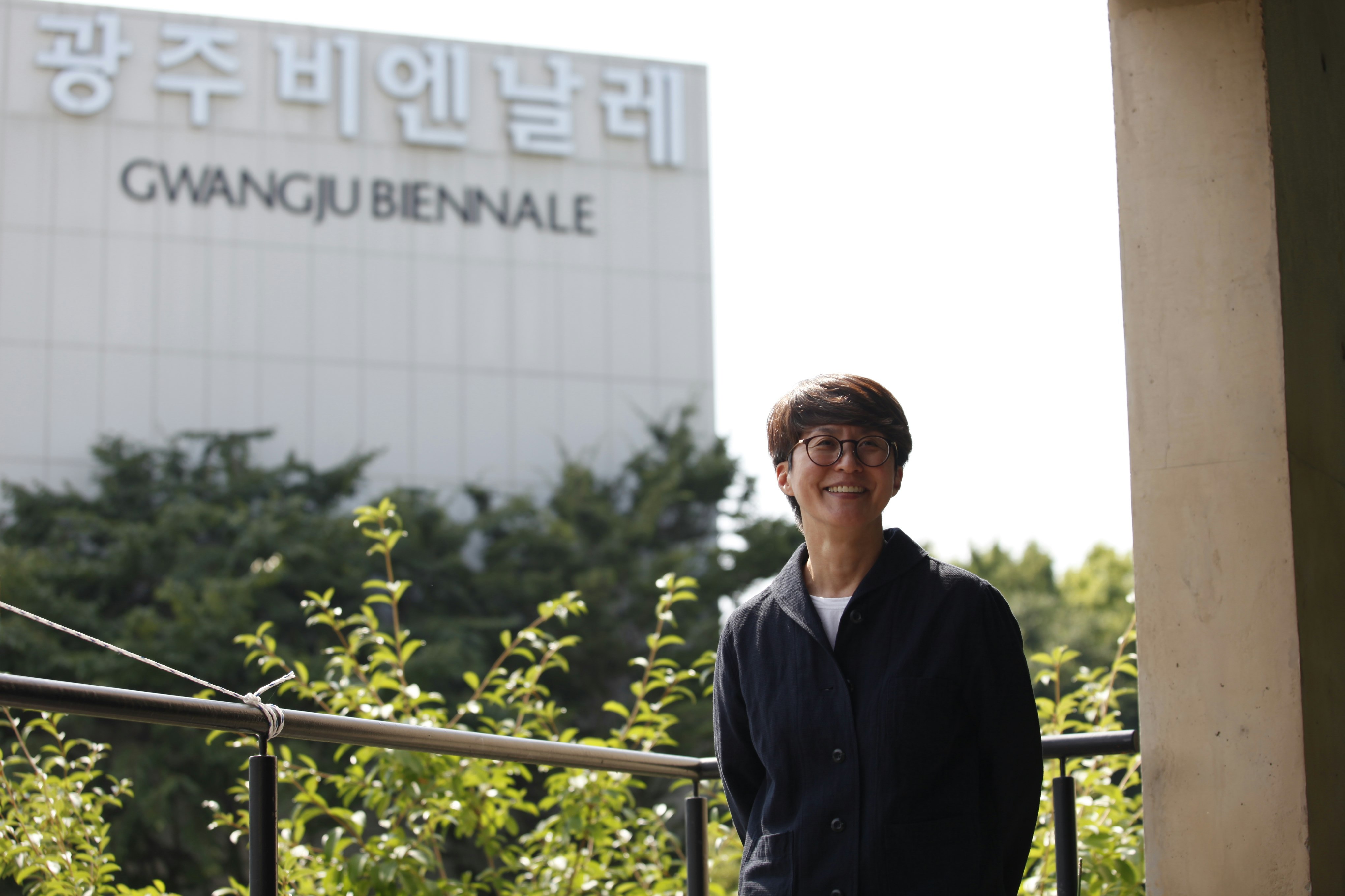 A South Korean curator with a short haircut smiles in front of a large building with a sign that reads, 'Gwangju Biennale', in English letters and Hangul.