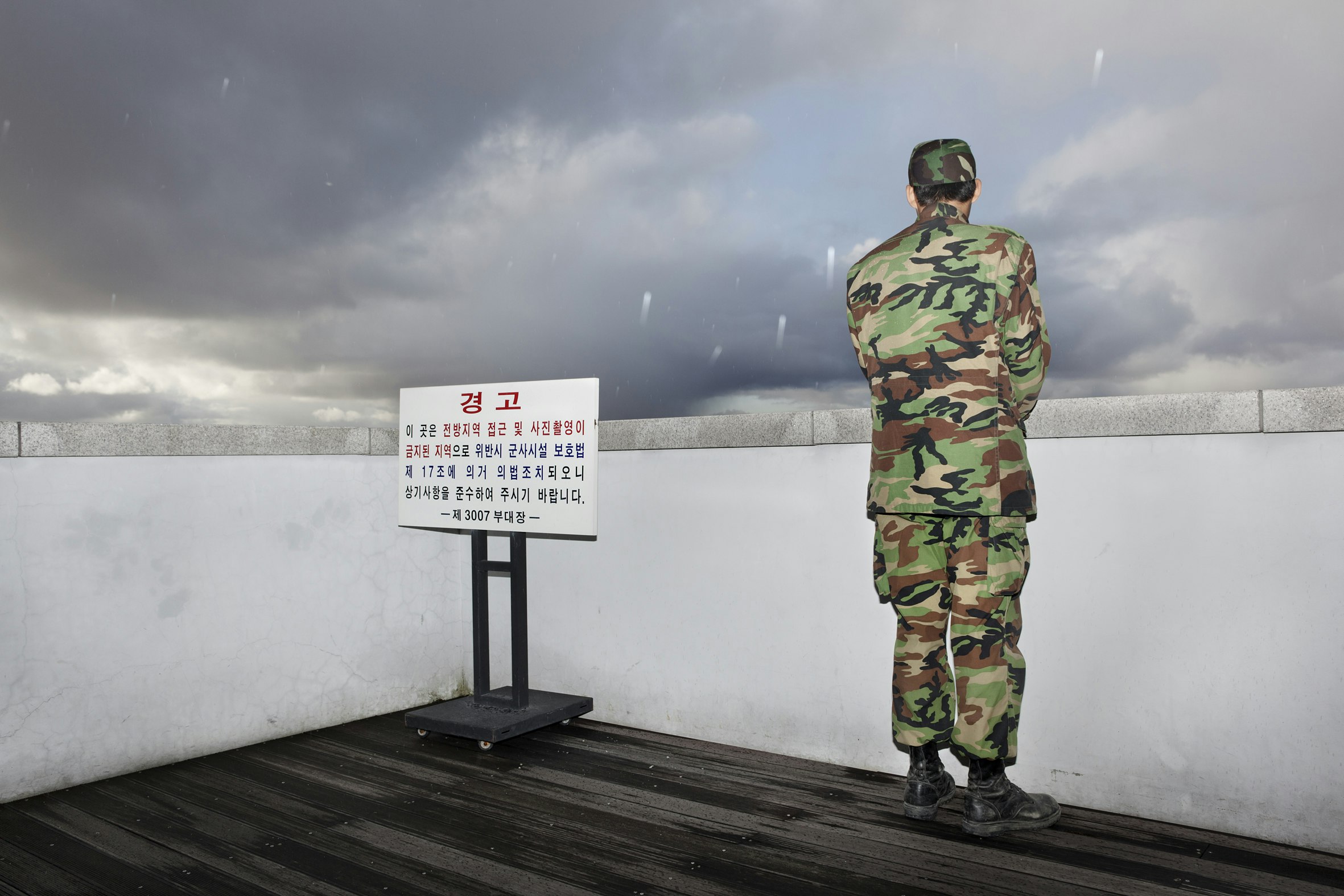 A painting of a soldier in camouflage and combat boots standing with his back to us, looking over a concrete landing as it snows outside.