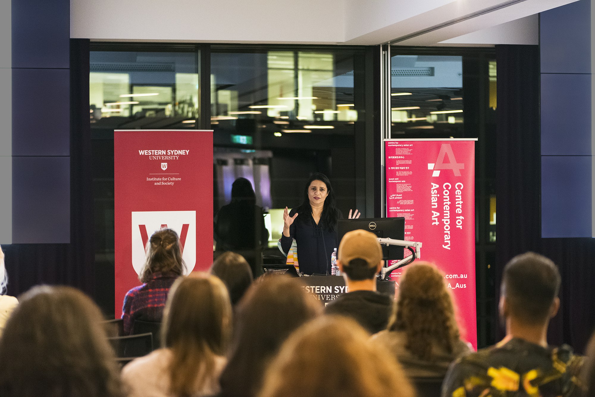 A female-presenting speaker with long black hair gestures to an audience sitting in front of her. Behind her is a red banner reading 'Western Sydney University' and a pink banner reading '4A Centre for Contemporary Asian Art'.