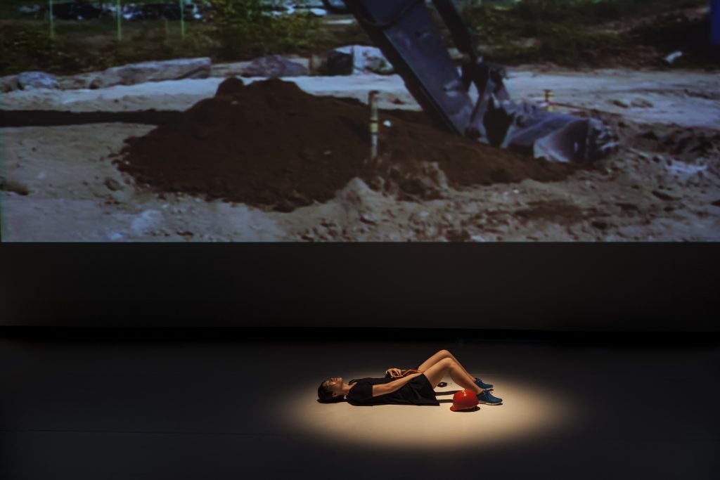 A Korean female-presenting figure in a short black dress lays on her back, under a spotlight, with her legs bent upright. Behind her is a projection of a mound of dirt at a construction site. 