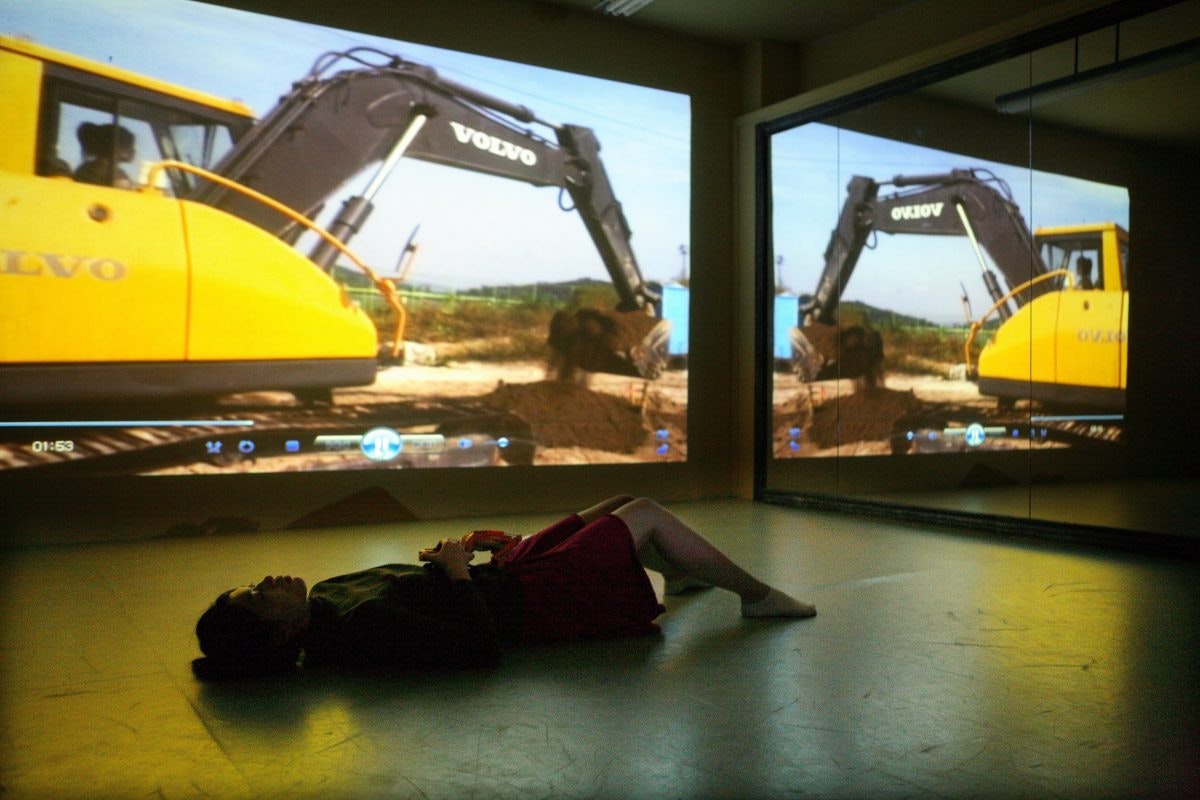 A Korean female-presenting figure lays on the ground in a dark exhibition space, while behind her is a projection of a bright yellow excavator picking up dirt.