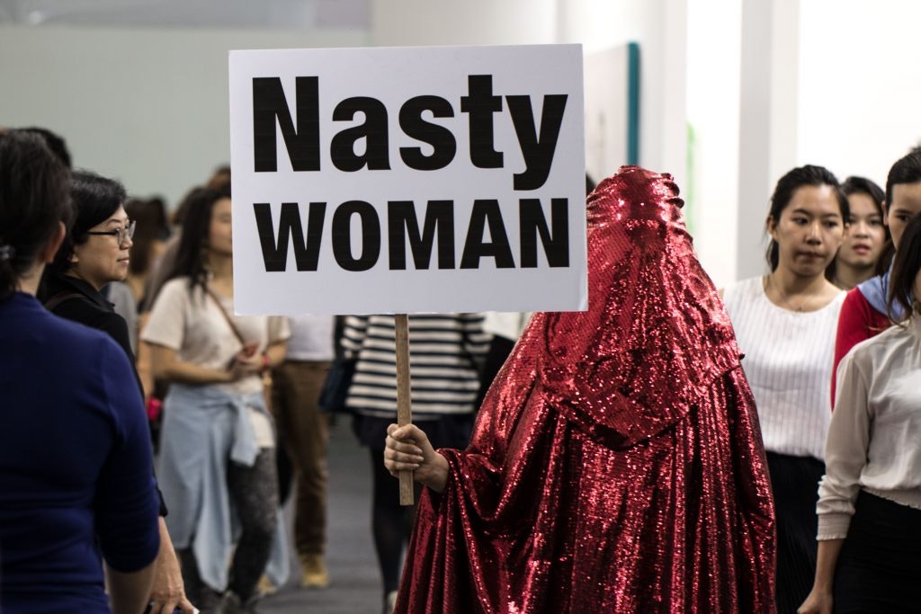 A figure wearing a sparkly red burqa stands with a placard that reads, 'Nasty WOMAN', while an art fair crowd walks around them.