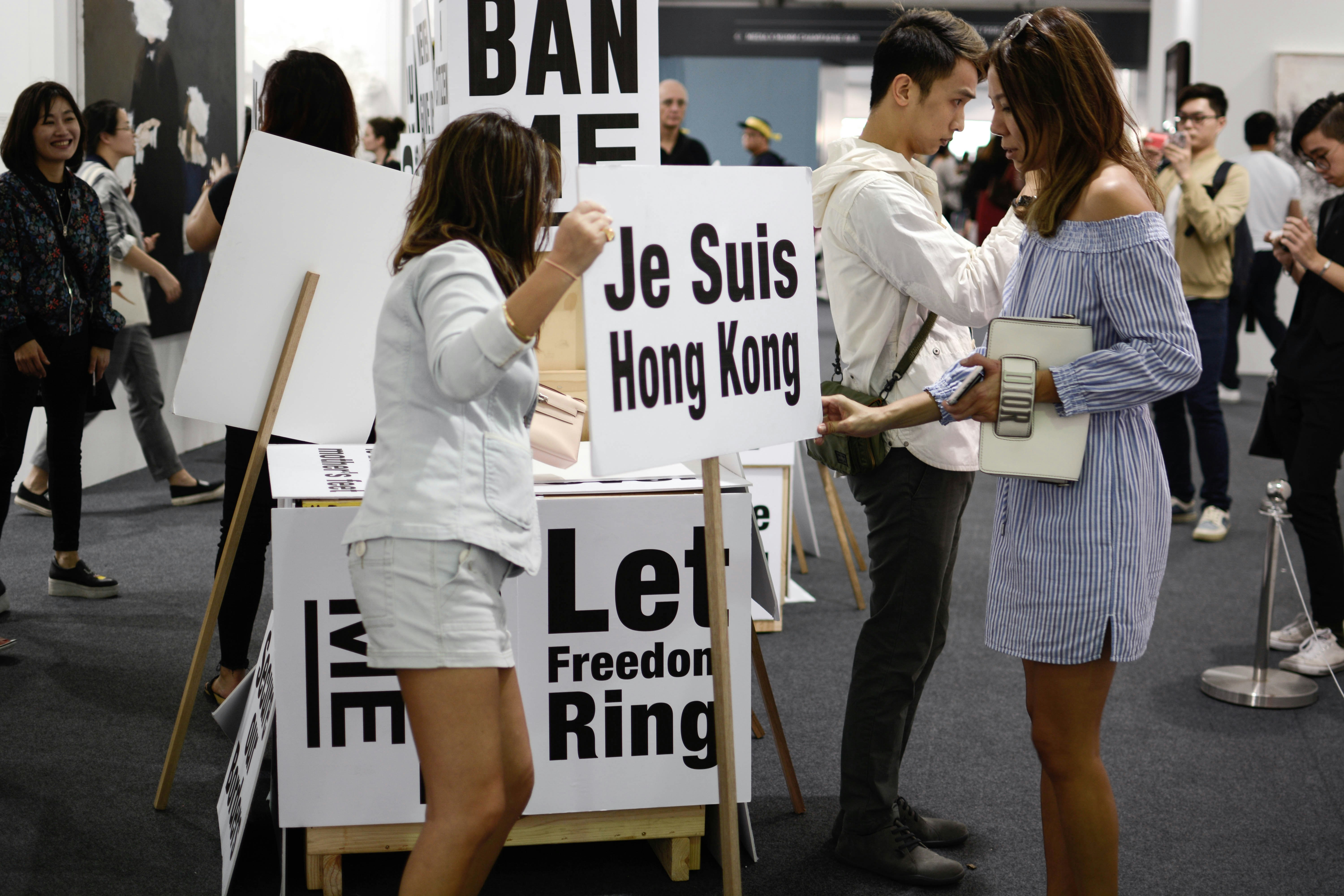 Two female presenting figures stand holding a placard that reads 'Je Suis Hong Kong'. A placard nearby reads, 'Let Freedom Ring'