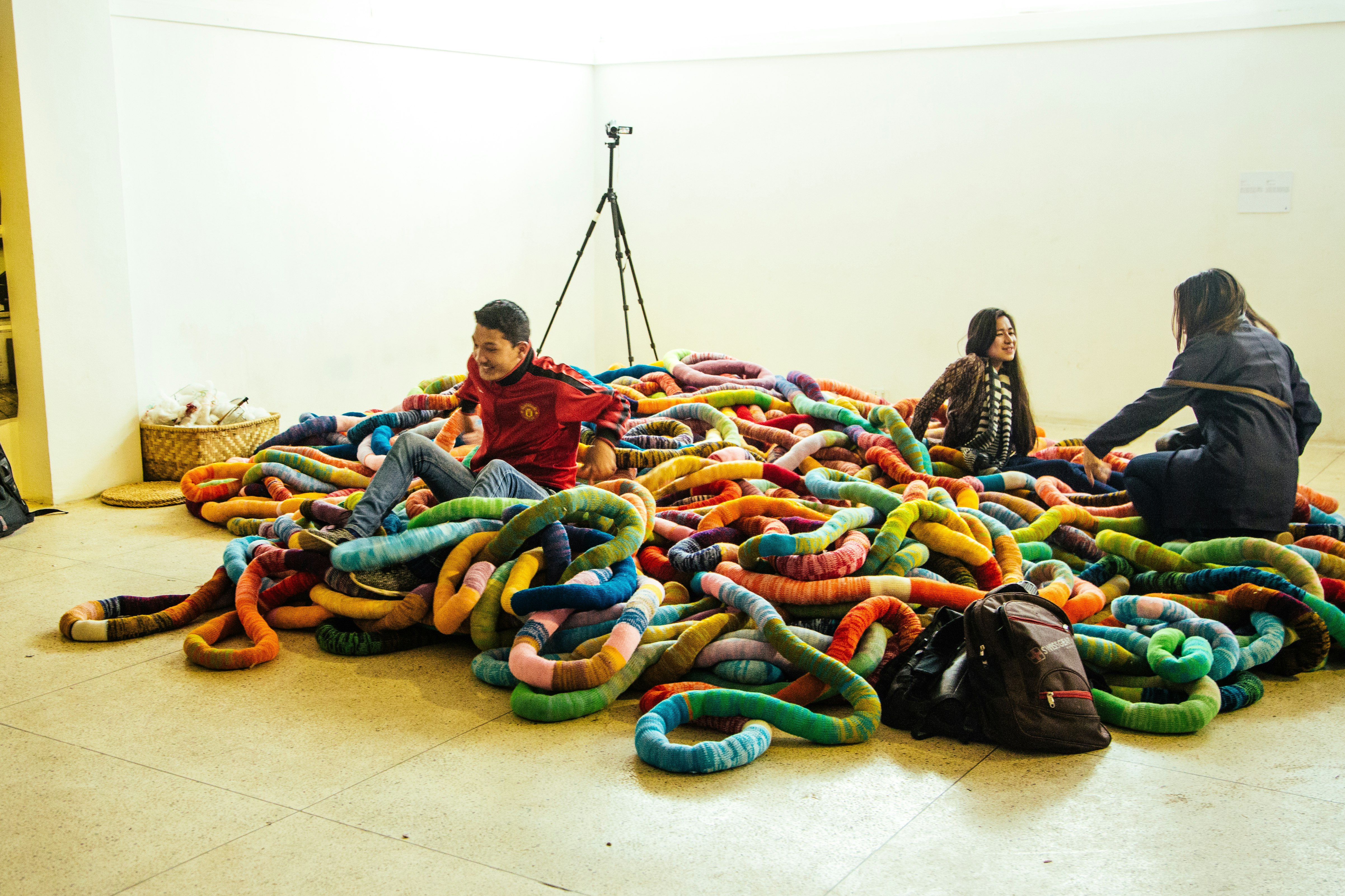 A smiling male-presenting figure and two female-presenting figures sit in a pile of woollen worm-like sculptures.
