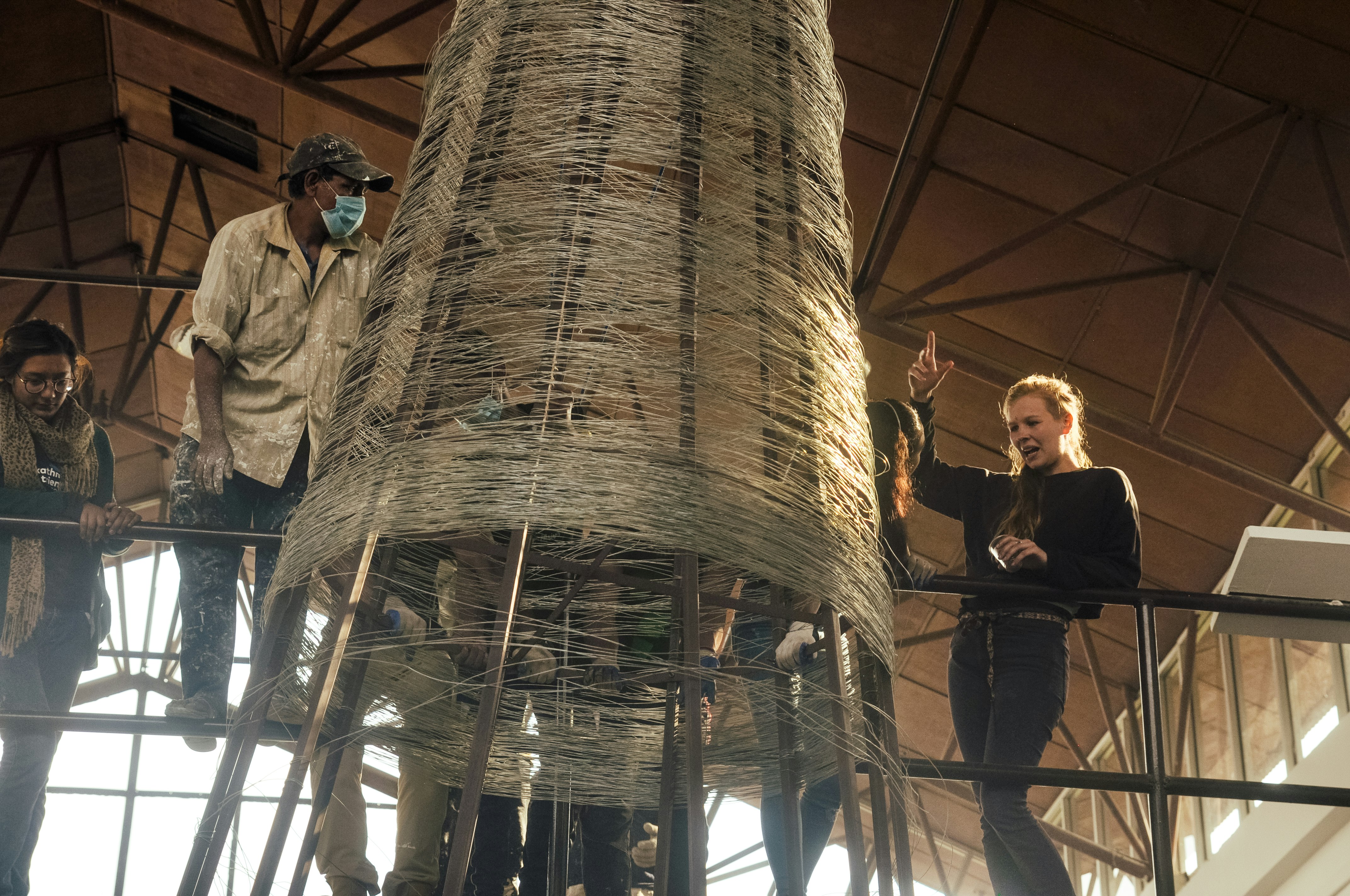 An installation team stand on a metal balcony, facing a immense cylindrical structure wrapped in metal.