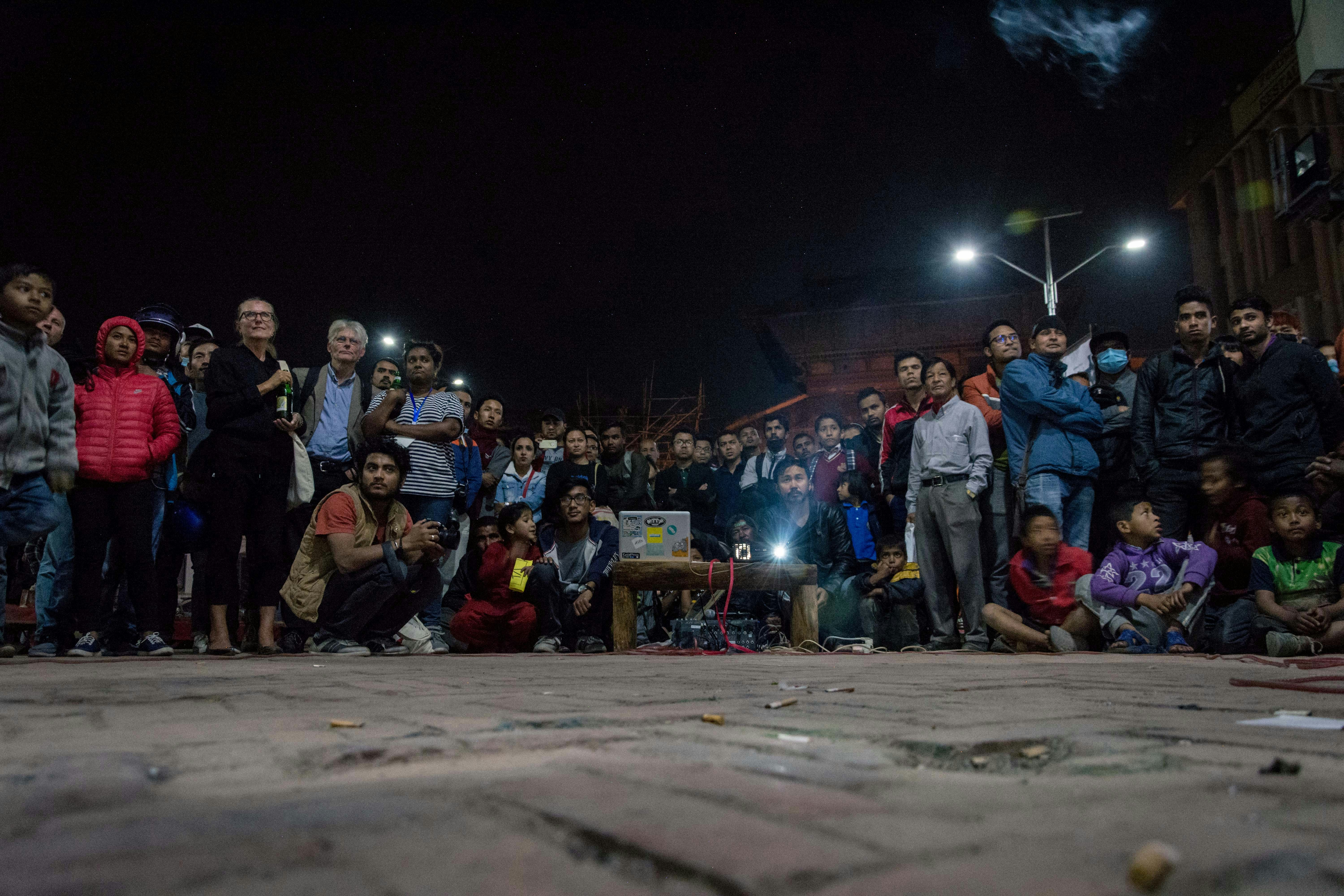 An audience gather outdoors at night at a screening, facing a projection of a short film.