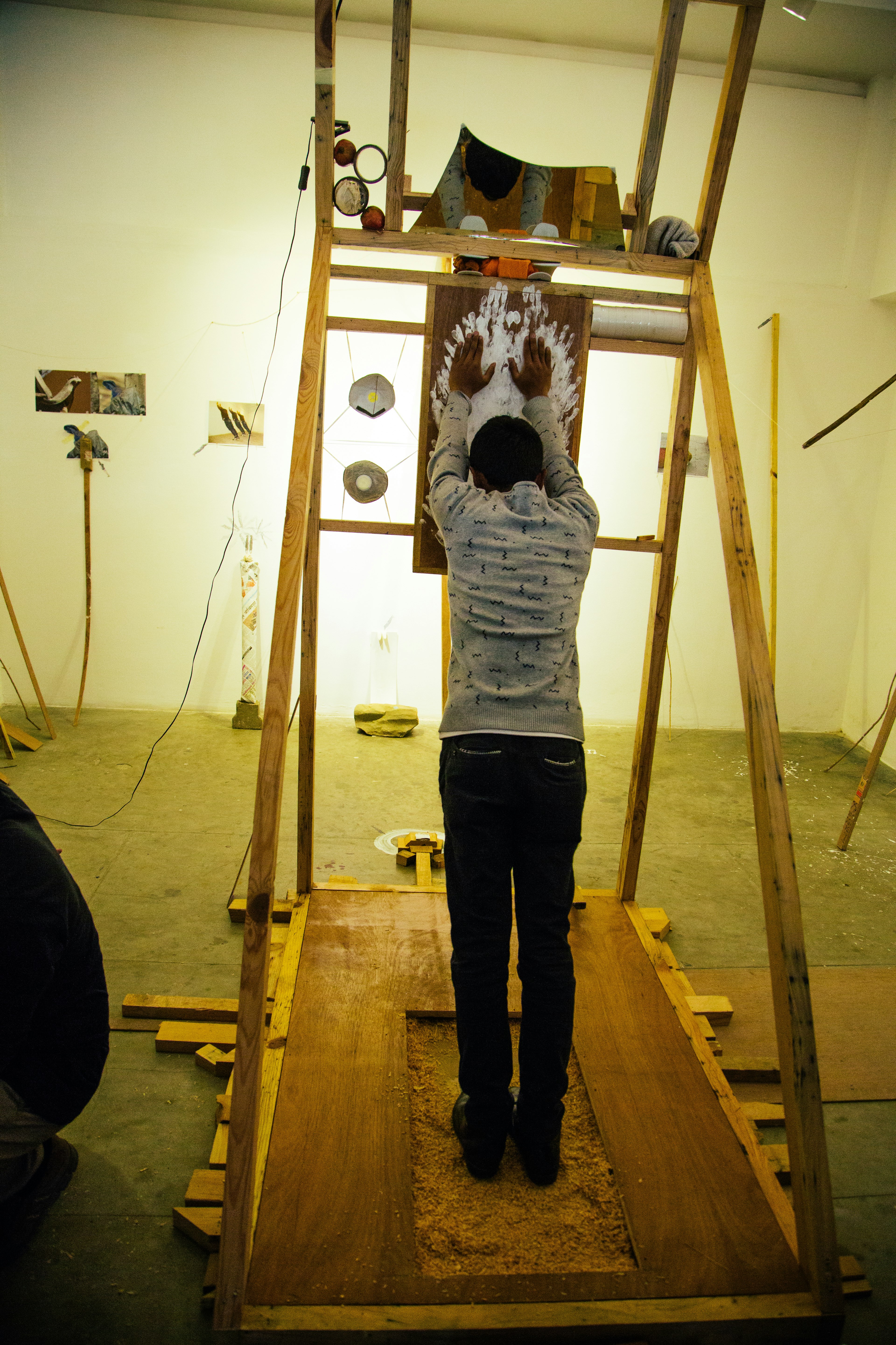 A male-presenting figure stands on a sloped wooden structure with their hands on a suspended wooden board that is painted with white handprints.