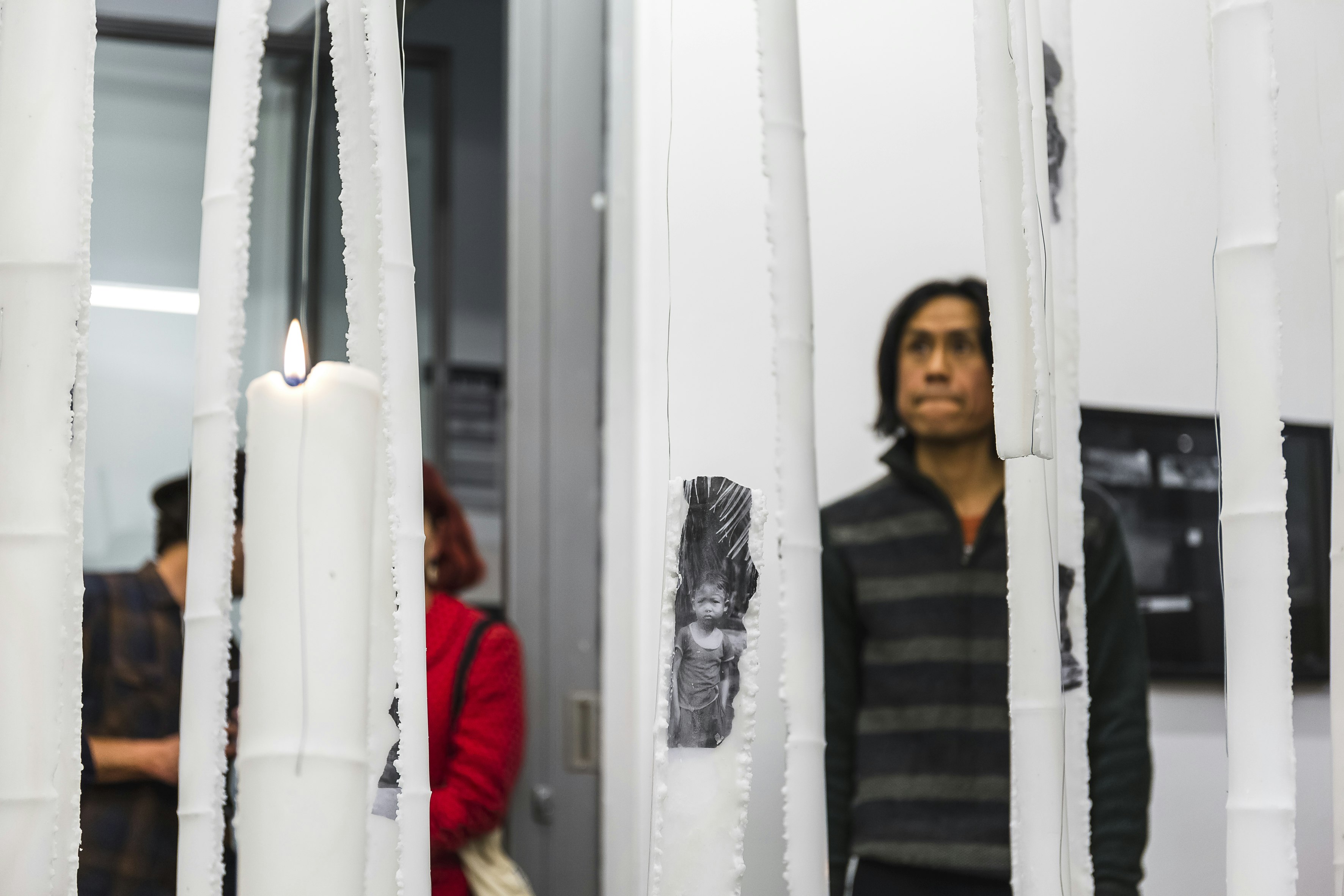 Dacchi stands before his installation in the ground floor 4A gallery space. A series of white candles and a small black-and-white photographic print of a boy is shown as part of the installation.