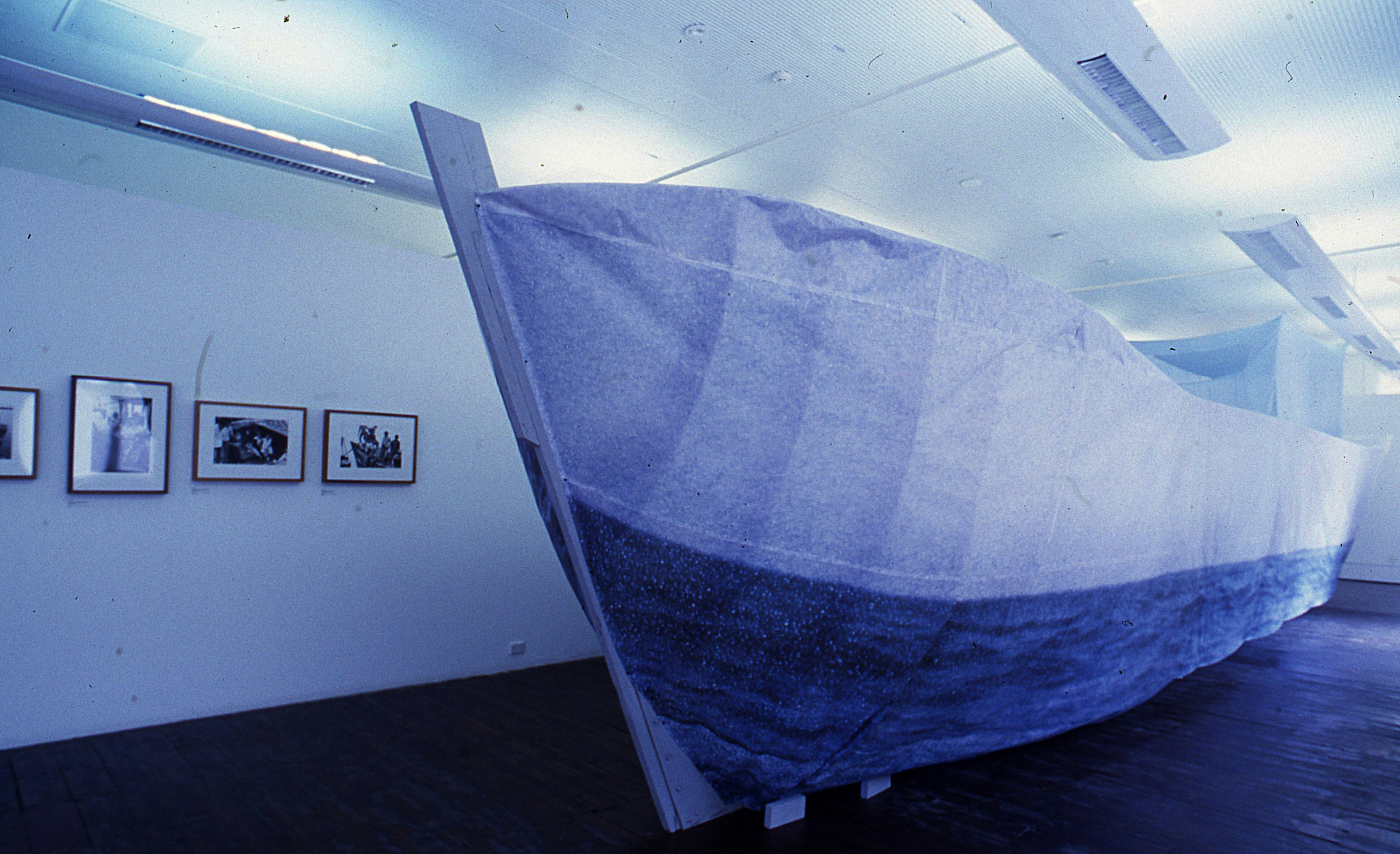 A large boat sculpture in a gallery space. On the wall nearby are framed black-and-white photographs.