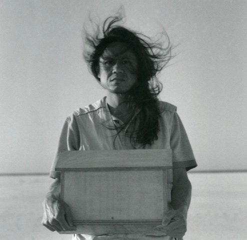 Dacchi stands holding a box and looking directly at the camera with a firm gaze. His hair, which reaches below his shoulders, blows in the wind.