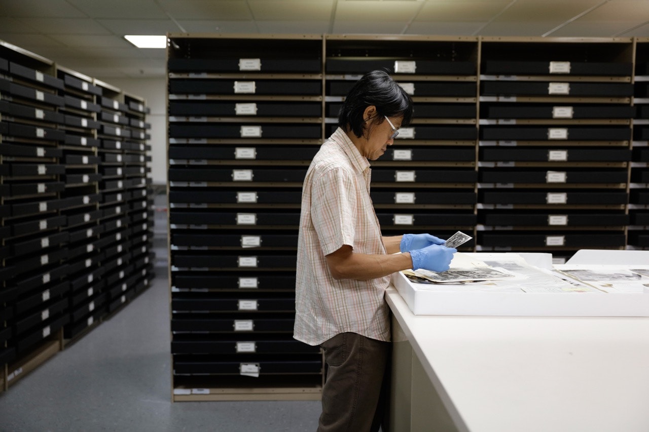 Dacchi stands in an archival room wearing blue protective gloves and glasses, looking at a series of black-and-white photographs.