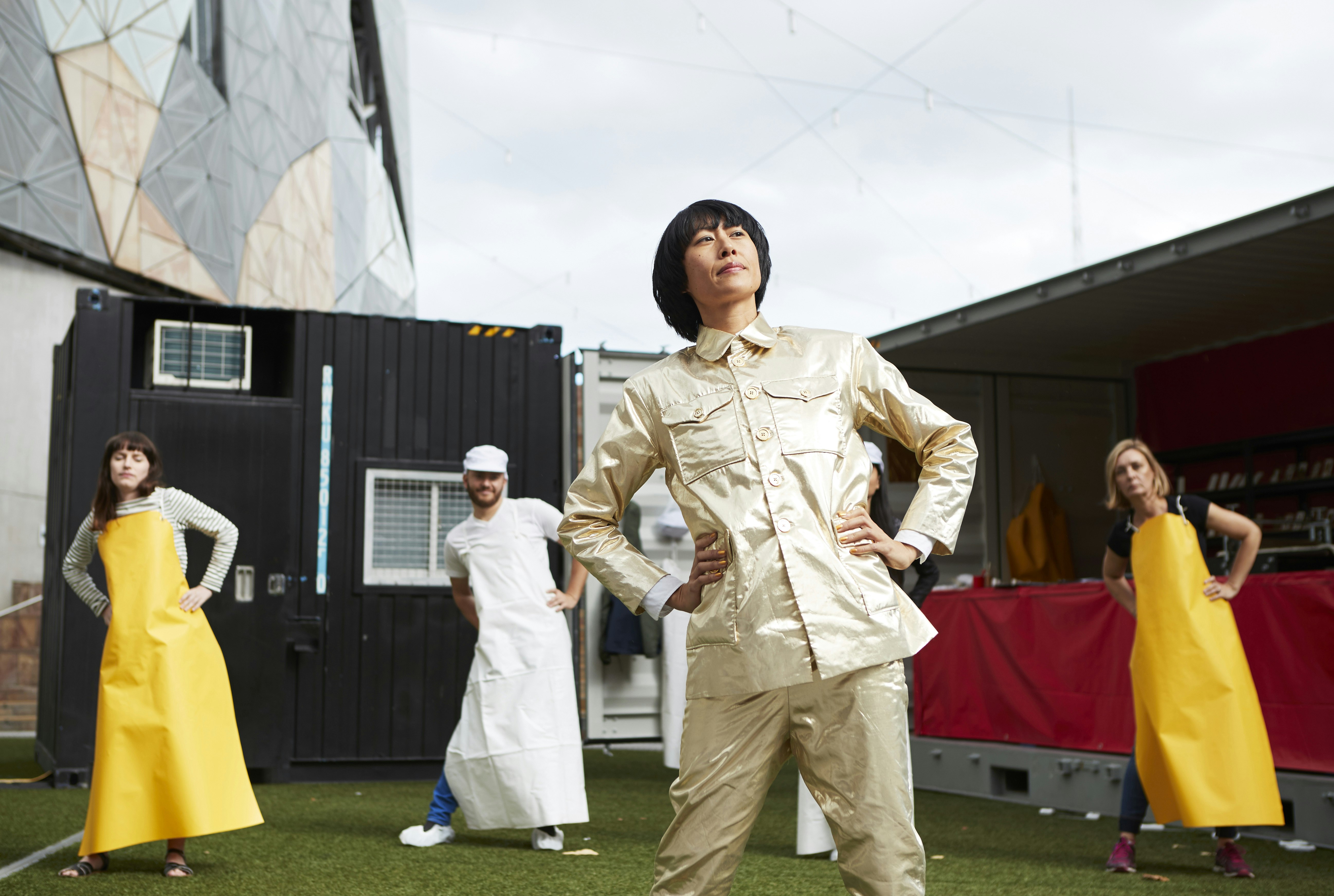 A female-presenting artist dressed in a gold suit stands with their hands on their hips, gazing off camera. Behind them is a line of 'workers' dressed in aprons, with their hands on their hips.