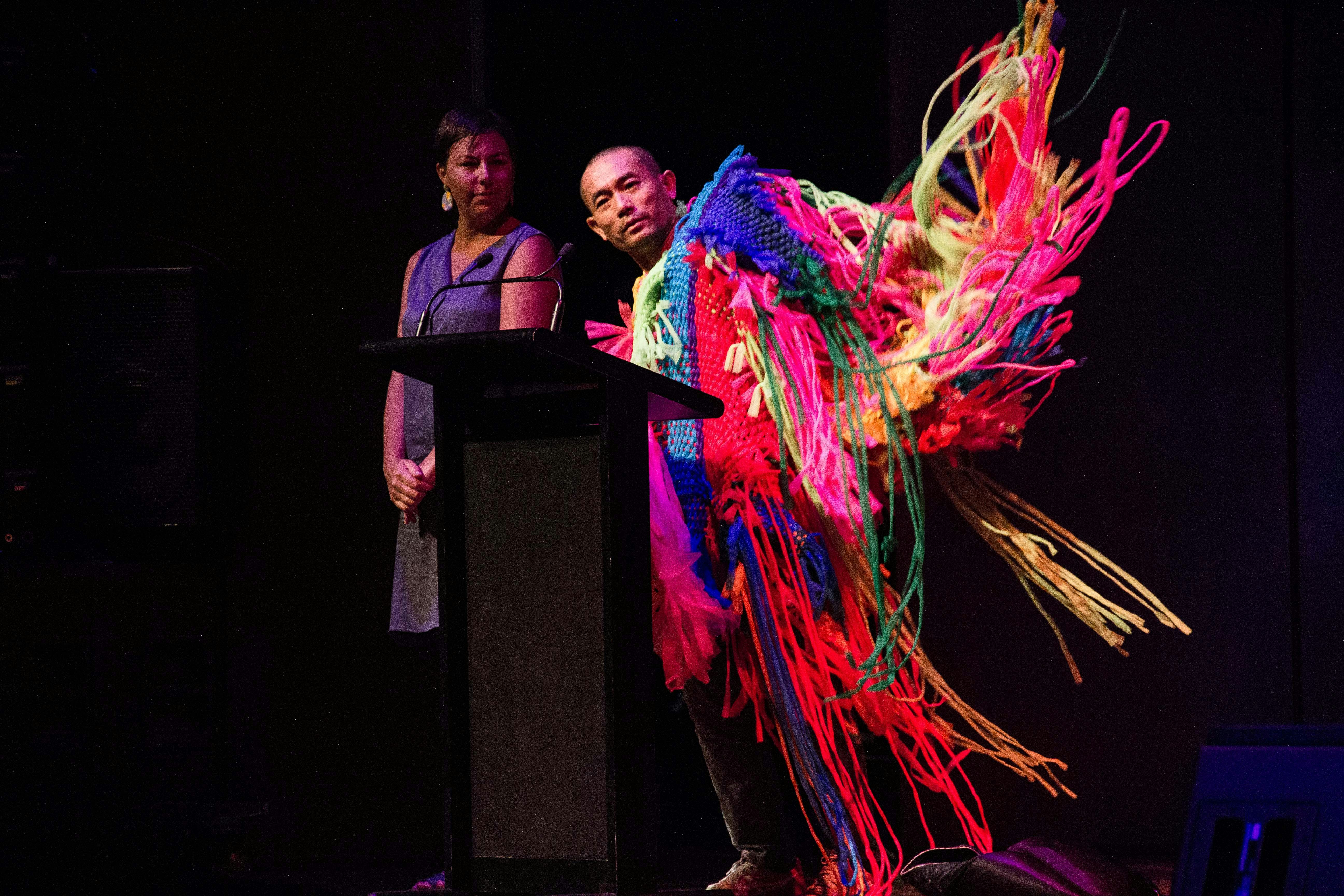 A male-presenting dancer in a flamboyant woven costume with draping threads dances at a lecturn in a dark auditorium.