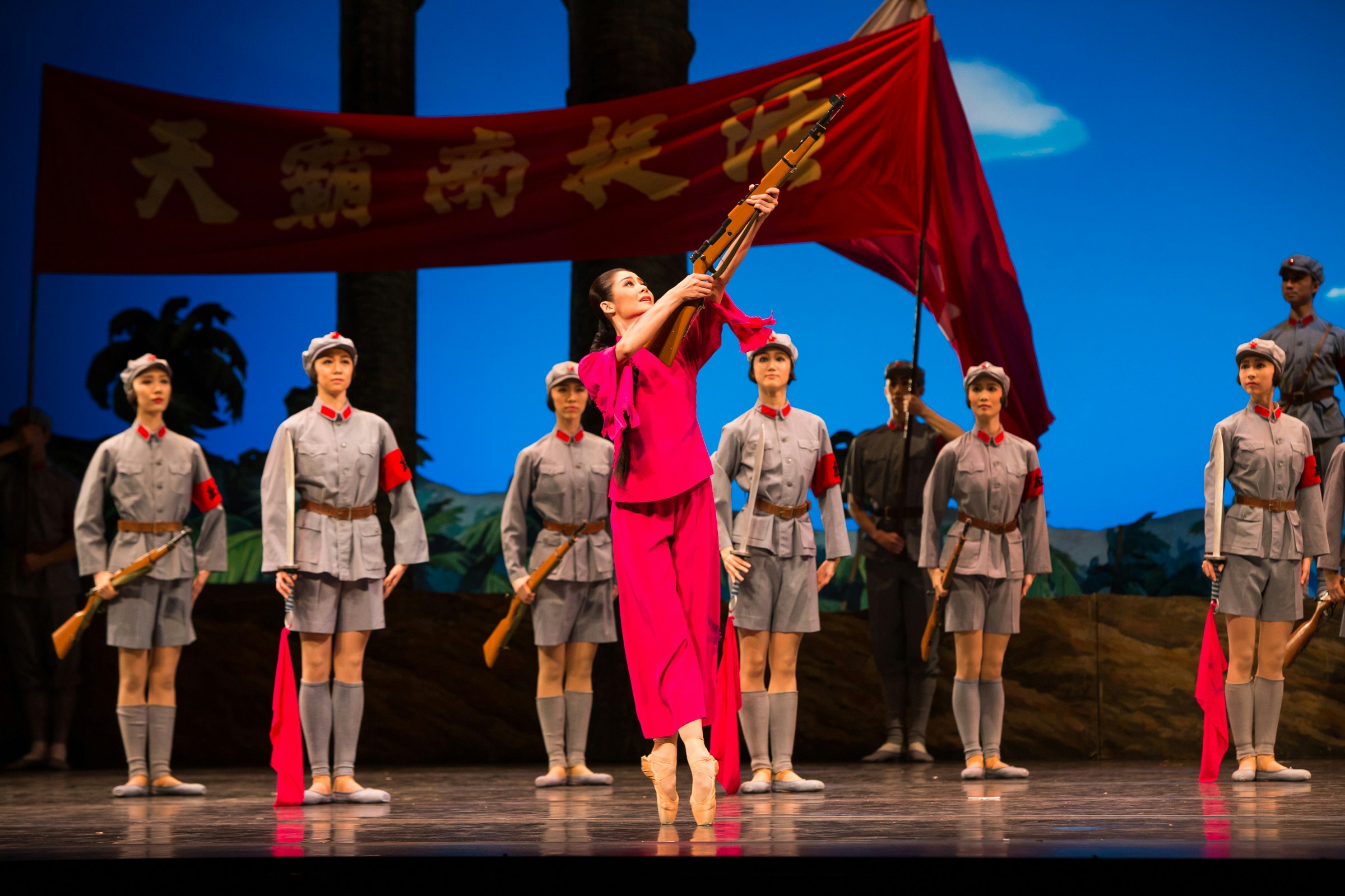 A Han Chinese female-presenting ballet dancer holds a rifle on stage. A line of ballet dancers dressed as Communist soldiers stand behind under a red banner painted with Chinese characters.