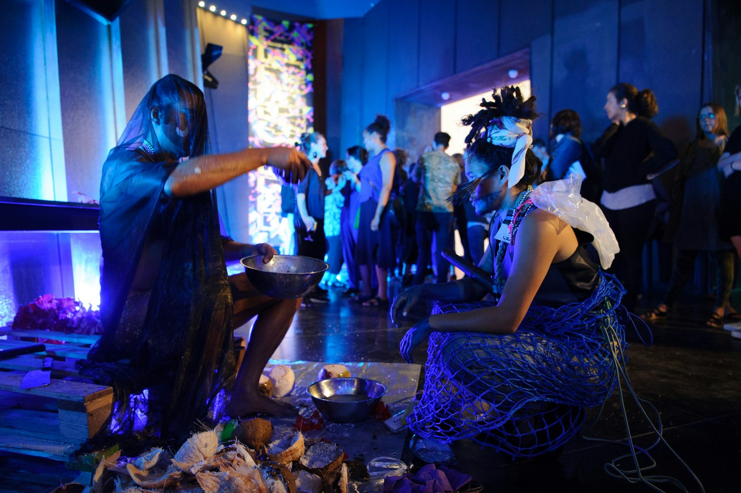 Two artists draped in ocean debris, netting and mesh sit facing each other in a blue-lit room.