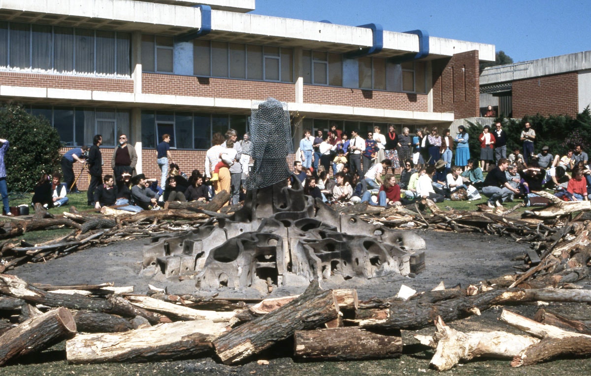 A burnt base of a sculptural tree, covered with chicken wire, stands in the middle of a circle of woodfire. A crowd is standing and sitting behind the sculpture.