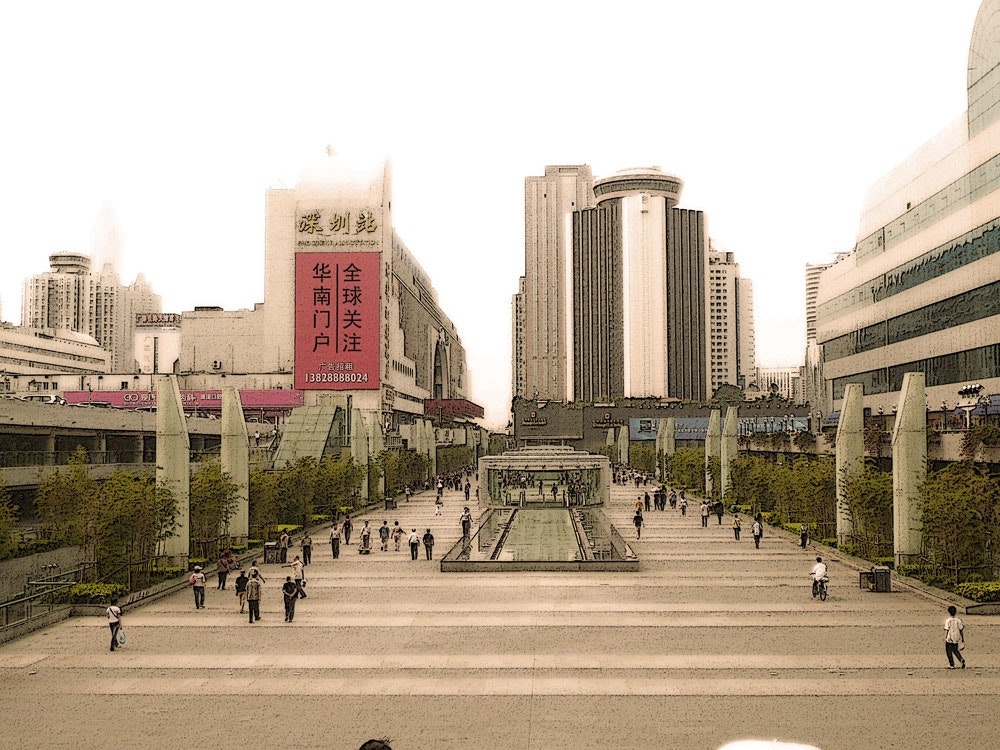 A long shot of Shenzhen city, populated with pedestrians and commuters on bikes. Skyscrapers and a red billboard populate the skyline.