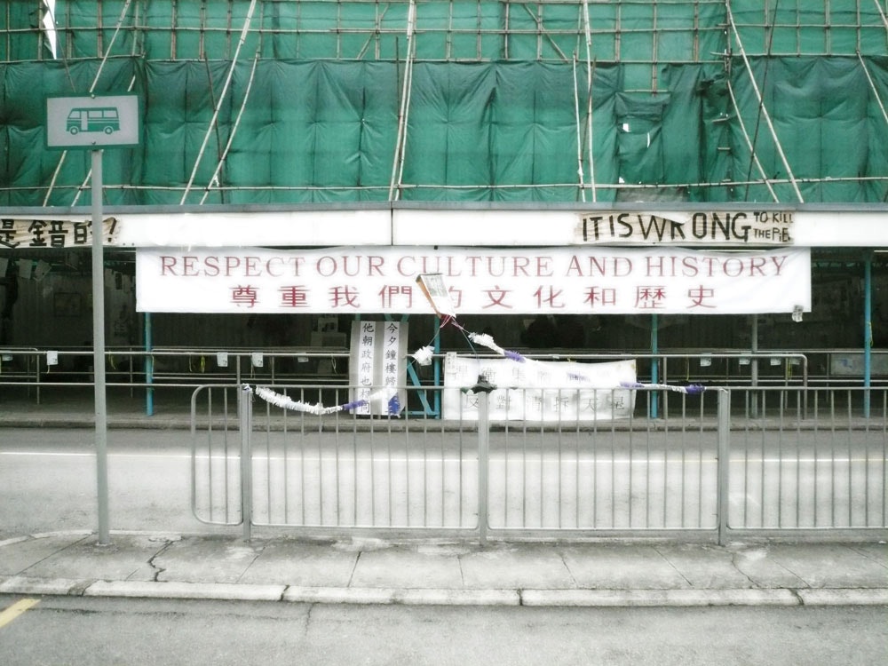 A shopfront with an awning that reads, Respect Our Culture and History, with Chinese characters underneath. A peeling sign above it reads, IT IS WRONG TO KILL THE PIER.