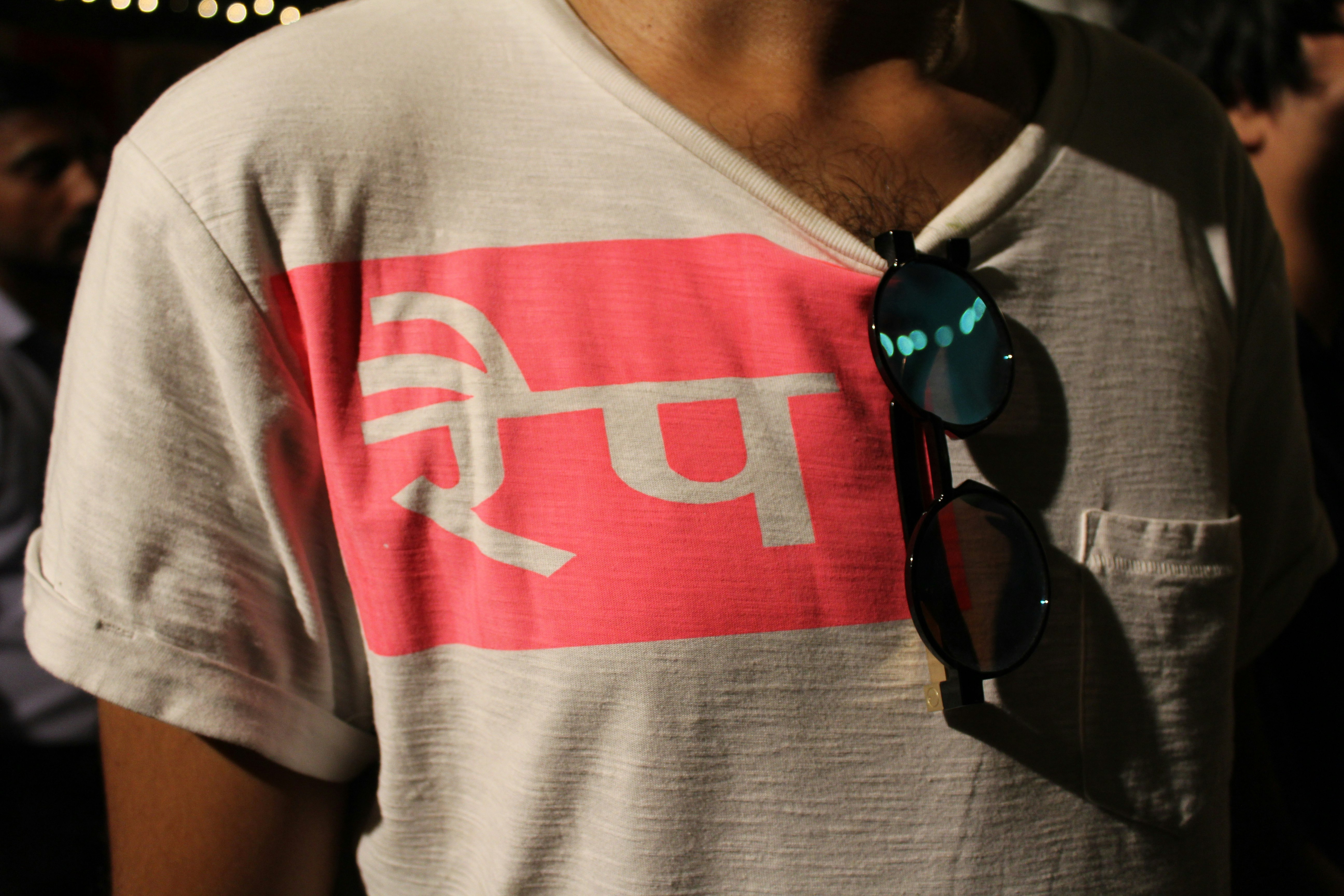 An anonymous figure wears a white t-shirt printed with Hindi characters in a bright pink square, with sunglasses resting on the collar of the t-shirt.