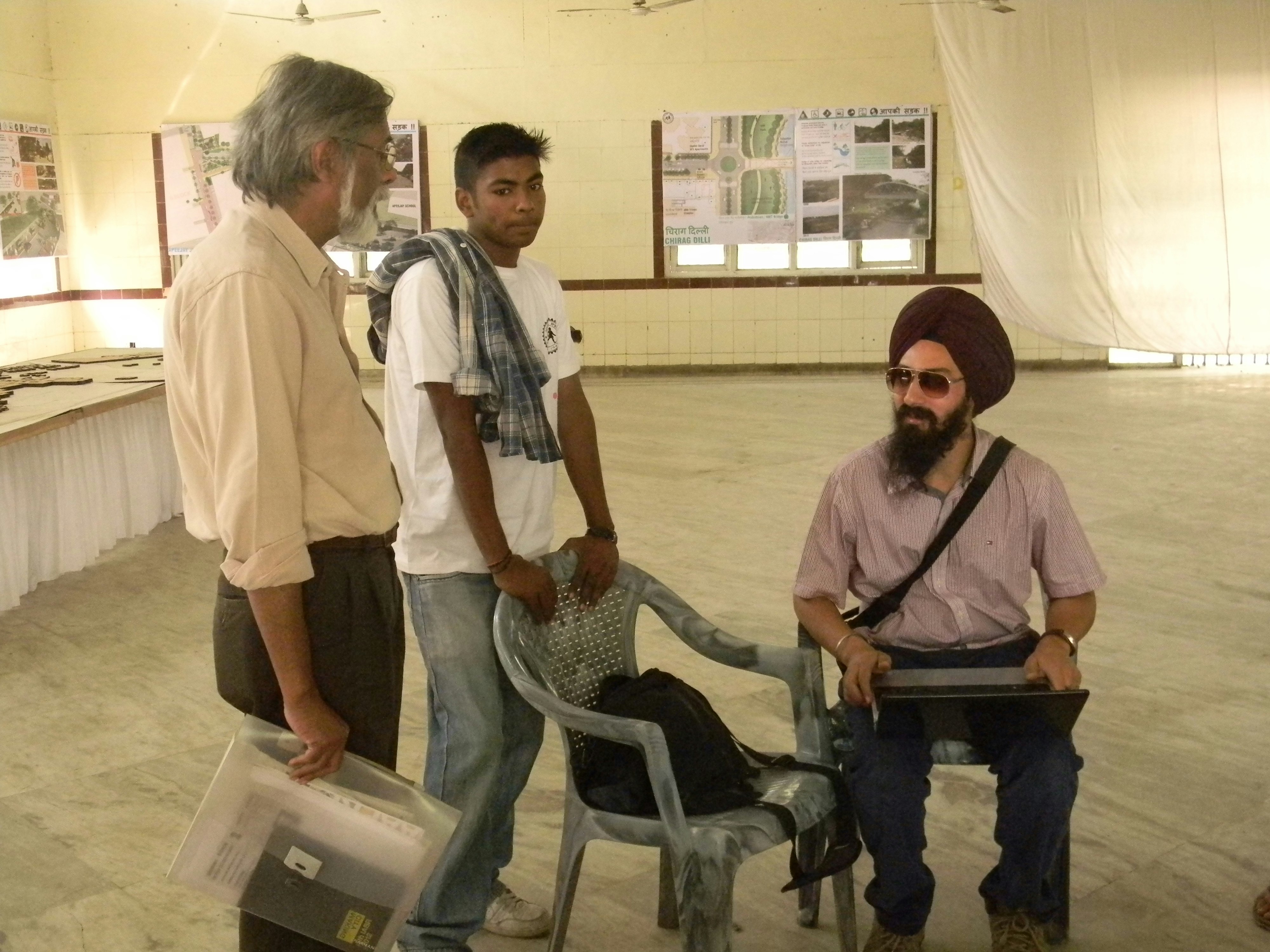 Three male-presenting figures stand in a room, speaking with each other. One of them has grey hair, wearing a dress shirt and trousers. The middle figure wears a white t-shirt and jeans. The third wears aviator sunglasses and a turban.