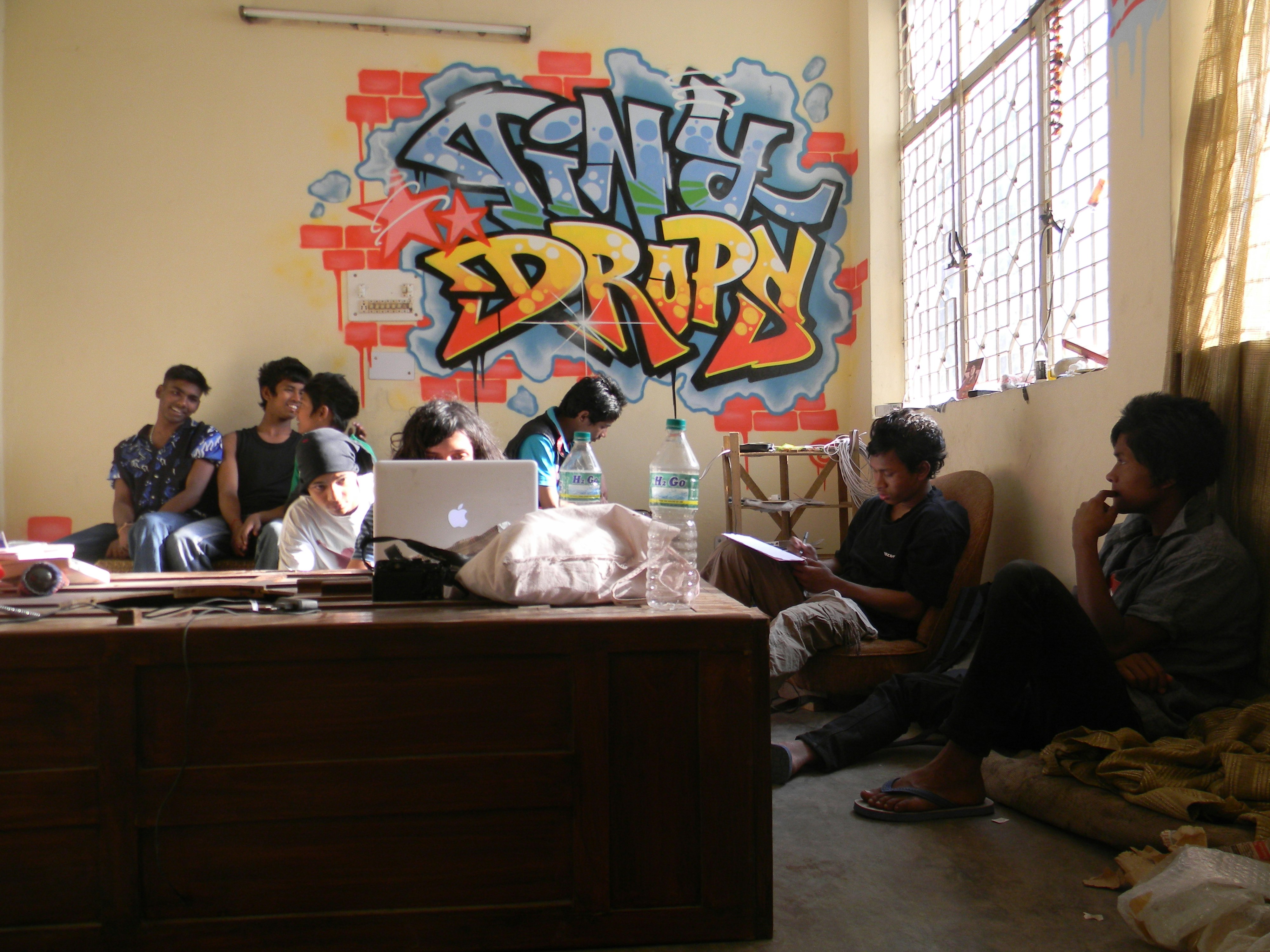 A group of male-presenting figures hang out in a room, some of them smiling at each other. TINY DROPS is graffitied in bright reds, yellows and blues on the wall behind them.