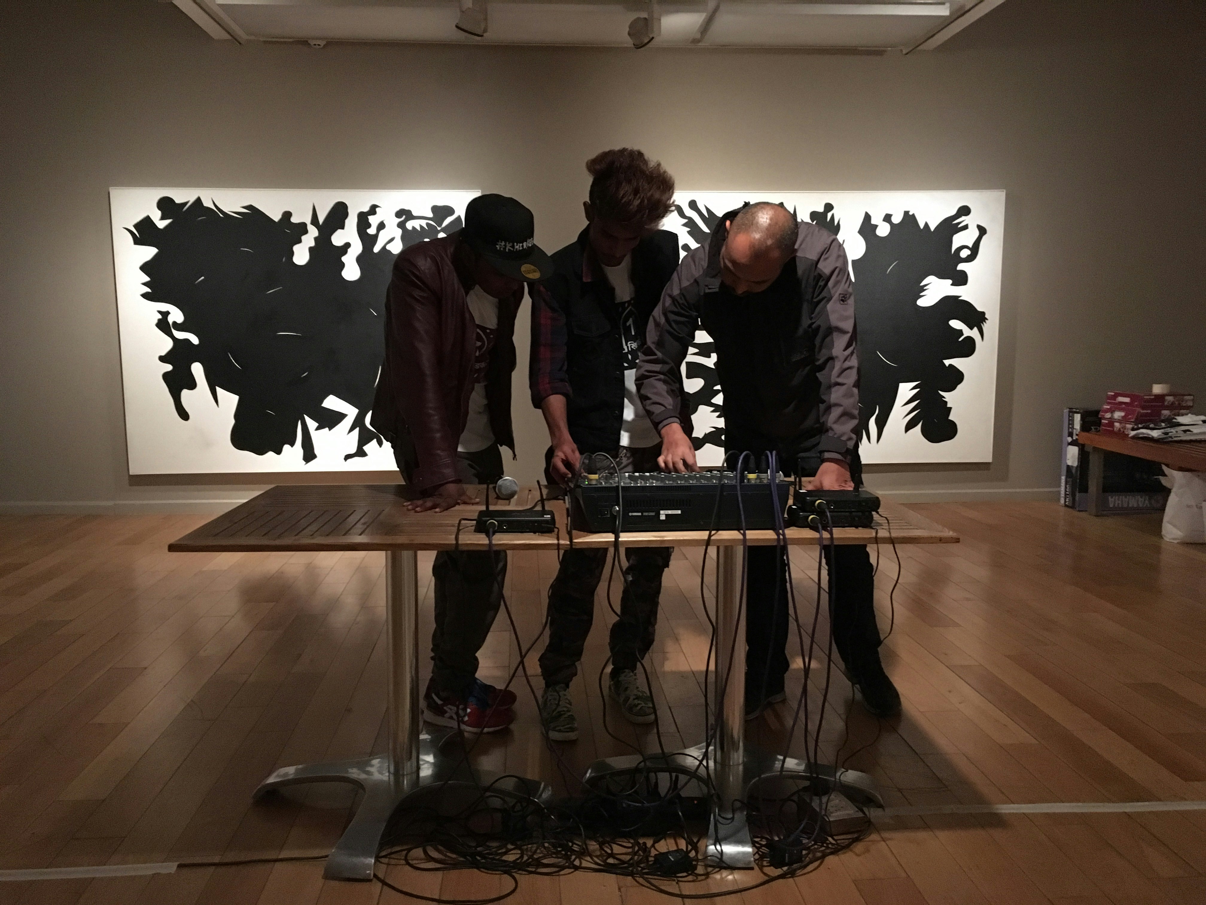 Three male-presenting figures stand in an art gallery, looking over a sound board. Behind them are two large white canvases painted with black shapes.