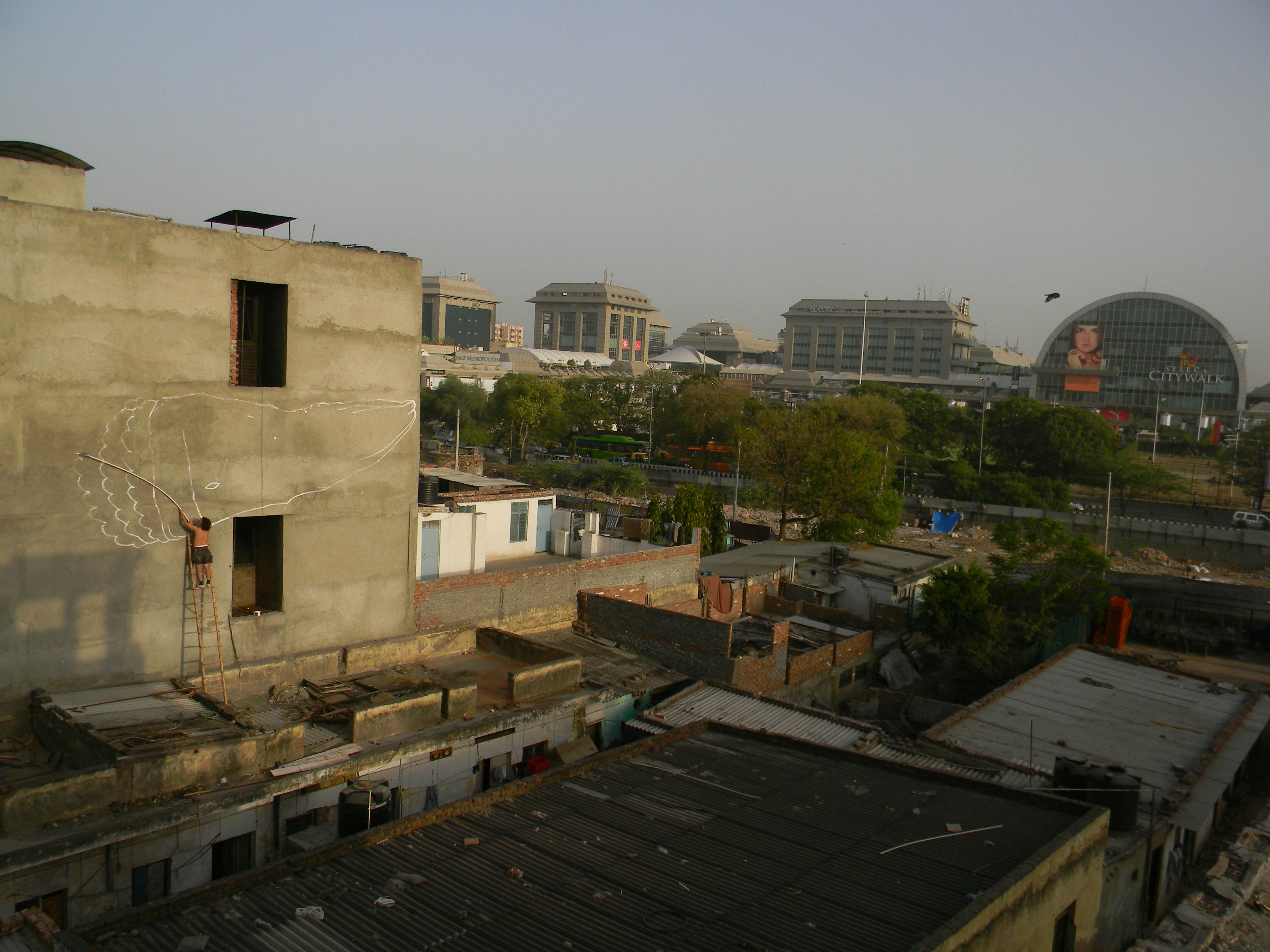 A view of some flat rooftops in Delhi. On the left is a figure standing on a stepladder, painting a large shape on a building facade.