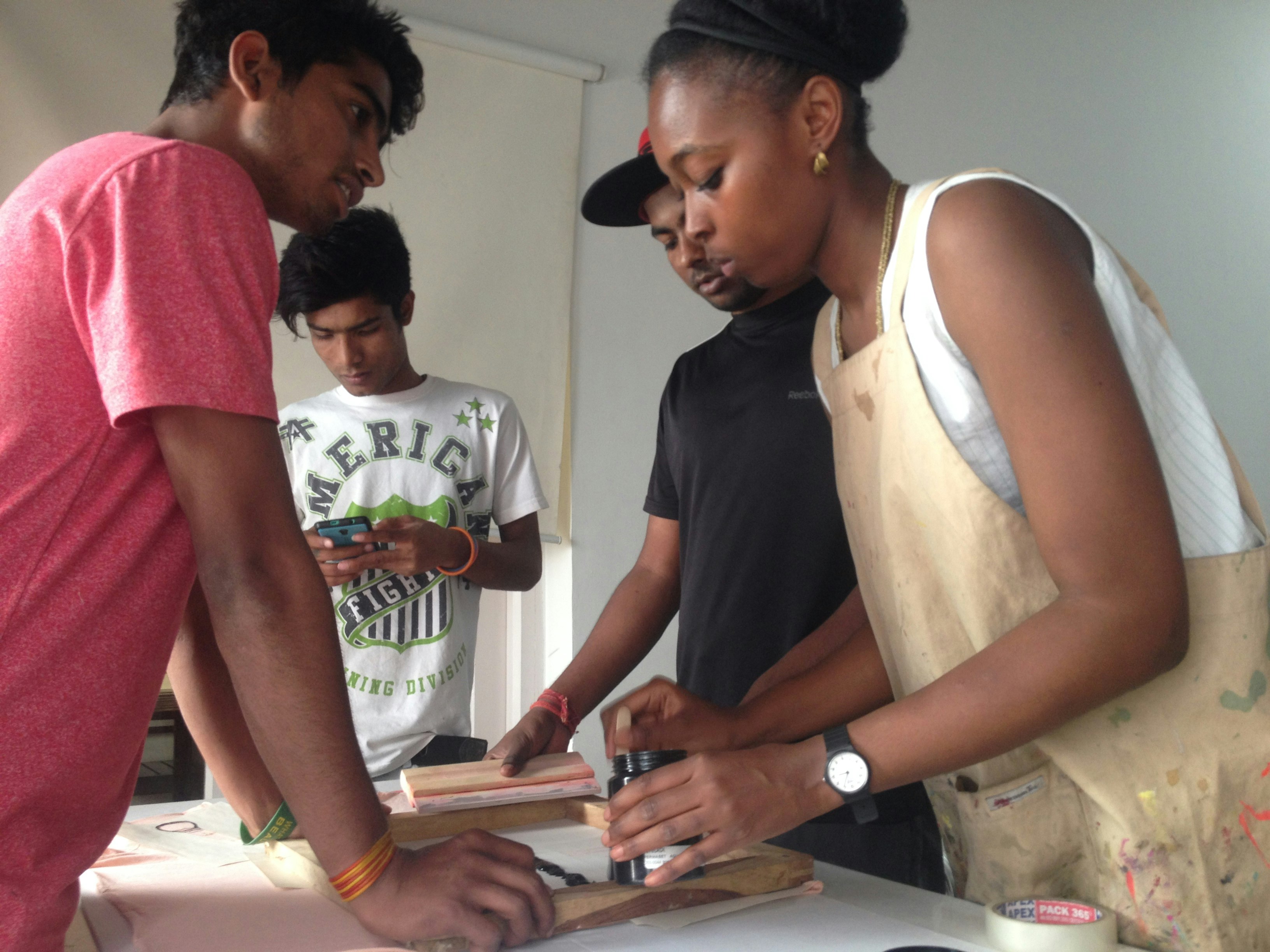 Yemi, a dark-skinned female-presenting artist wearing an artist's smock, shows three male-presenting figures how to screen print.