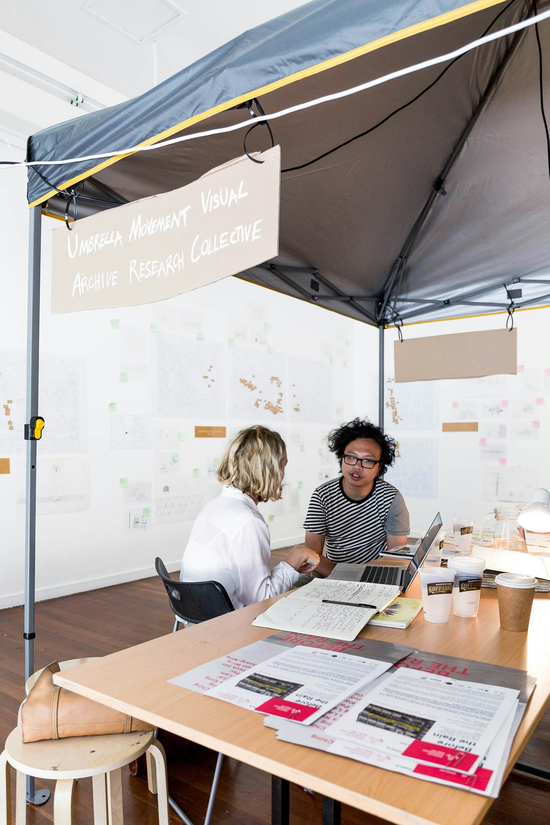Sampson Wong, an East Asian male-presenting figure in a black and white striped t-shirt and glasses, talks to a blonde female-presenting figure under a gazebo with the banner: Umbrella Movement Visual Archive Research Collective.