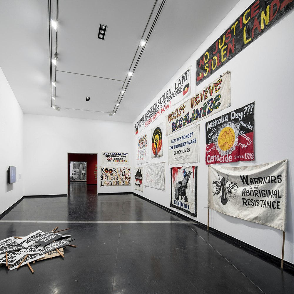 Protest banners in a white gallery space, with a pile of placards on the floor. Some of the banners read: WARRIORS OF THE ABORIGINAL RESISTANCE; NO JUSTICE ON STOLEN LAND; LEST WE FORGET THE FRONTIER WARS BLACK LIVES; GENOCIDE; RESIST REVIVE DECOLONIZE.