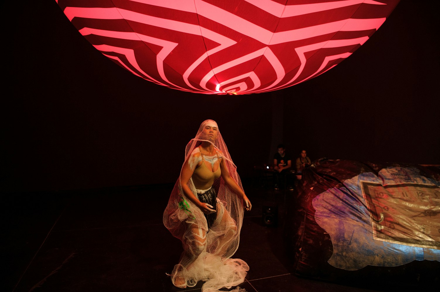A female-presenting dancer wearing a mesh veil, performing under a large pink striped lantern.