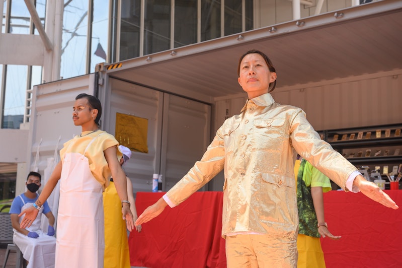 Eugenia and Jeremy, a Filipinx dancer wearing gold sparkly eyeshadow and a white apron, do calisthenics in front of a shipping container.