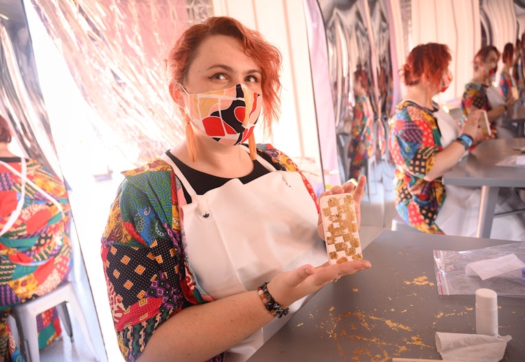 A Caucasian person with bright red hair wearing a face mask and a white apron holds up a gold phone.