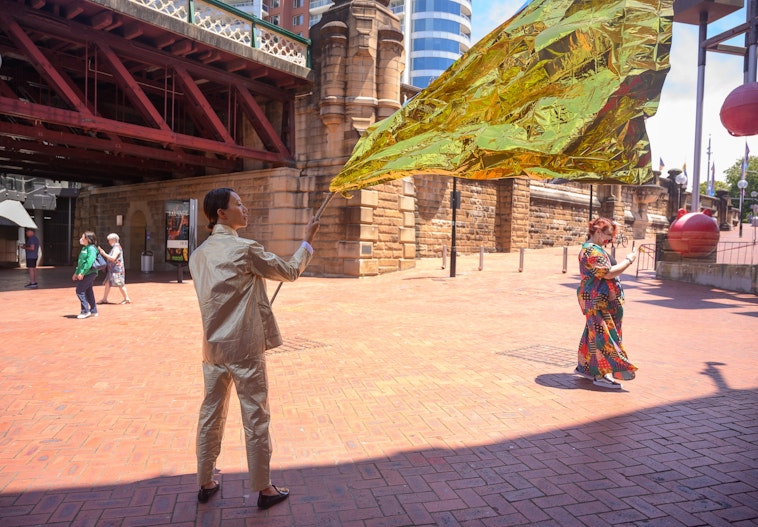 Eugenia, an East Asian woman wearing a two-piece gold suit, is flying a gold flag. 