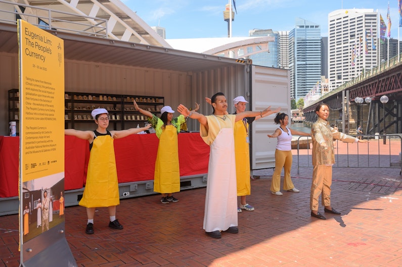 Performers dressed in yellow and white factory aprons stand with their arms outstretched in front of a shipping container.