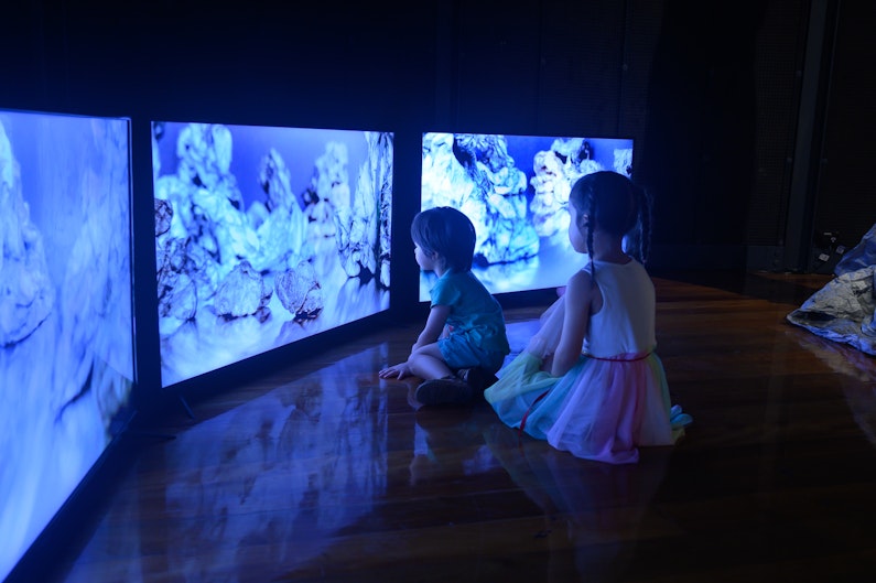 Two children sit on a dark gallery floor, in front of three television screens showing bundles of soft paper.