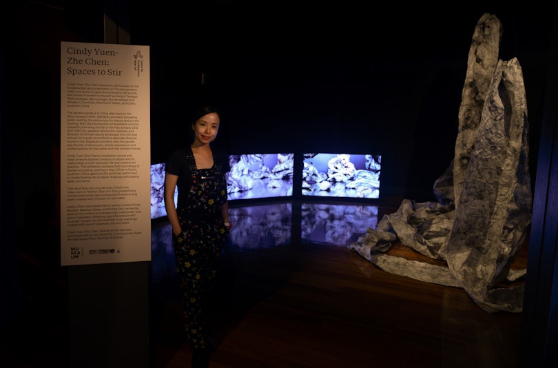 Cindy, an East Asian woman wearing a dark dress, stands inside a low-lit museum space next to a screen installation and a serie of paper scrolls suspended from the ceiling.