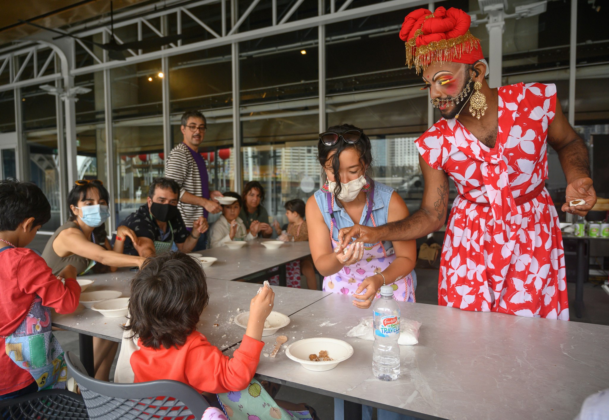 Radha, a South Asian drag performer, wears a red floral dress and a red headscarf while helping children make snacks.