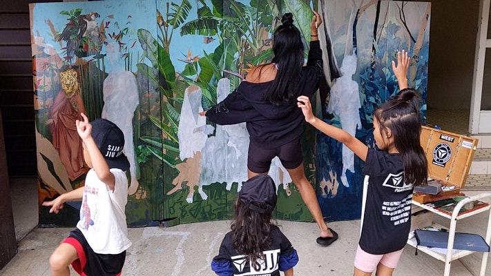 Marikit, a Filipina woman with long black hair, paints while her three children dance behind her.