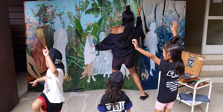 Marikit, a Filipina woman with long black hair, paints while her three children dance behind her.