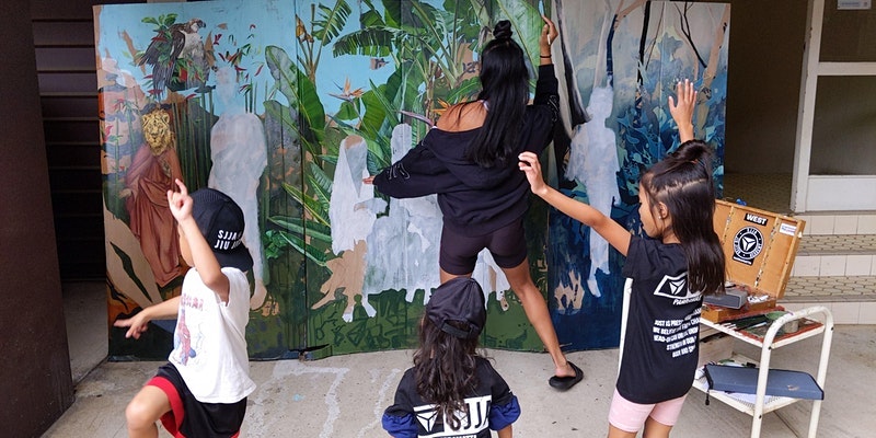 Marikit, a Filipina woman with long black hair, paints while her three children dance behind her.