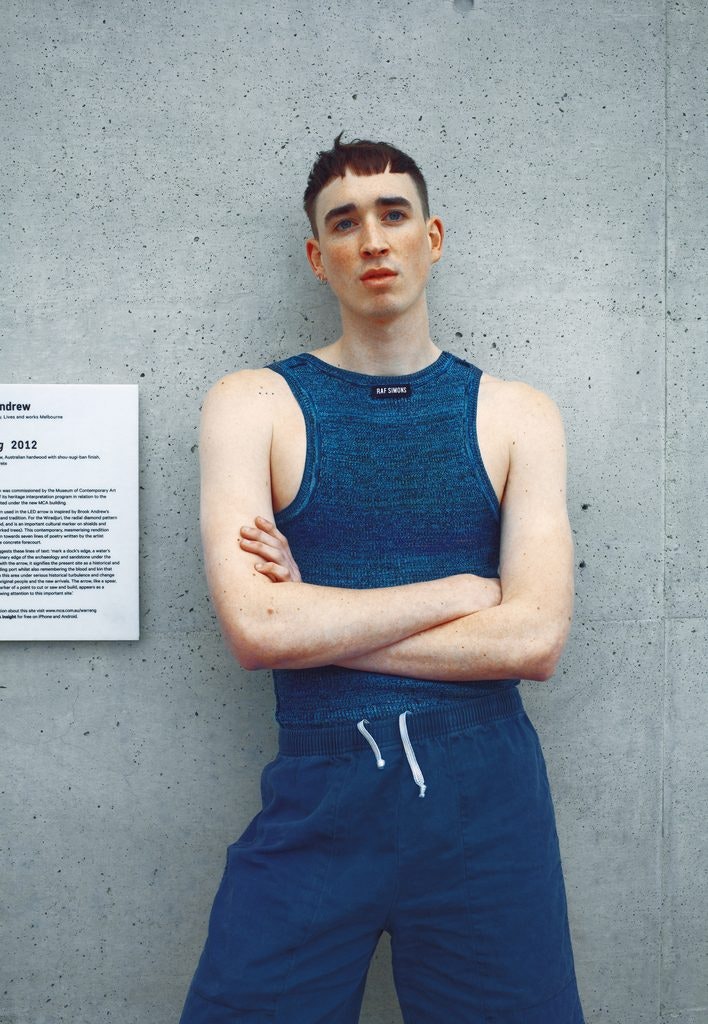 A Caucasian model with a baby fringe, wearing a blue knit tank top, stands with their arms crossed in front of a concrete wall.