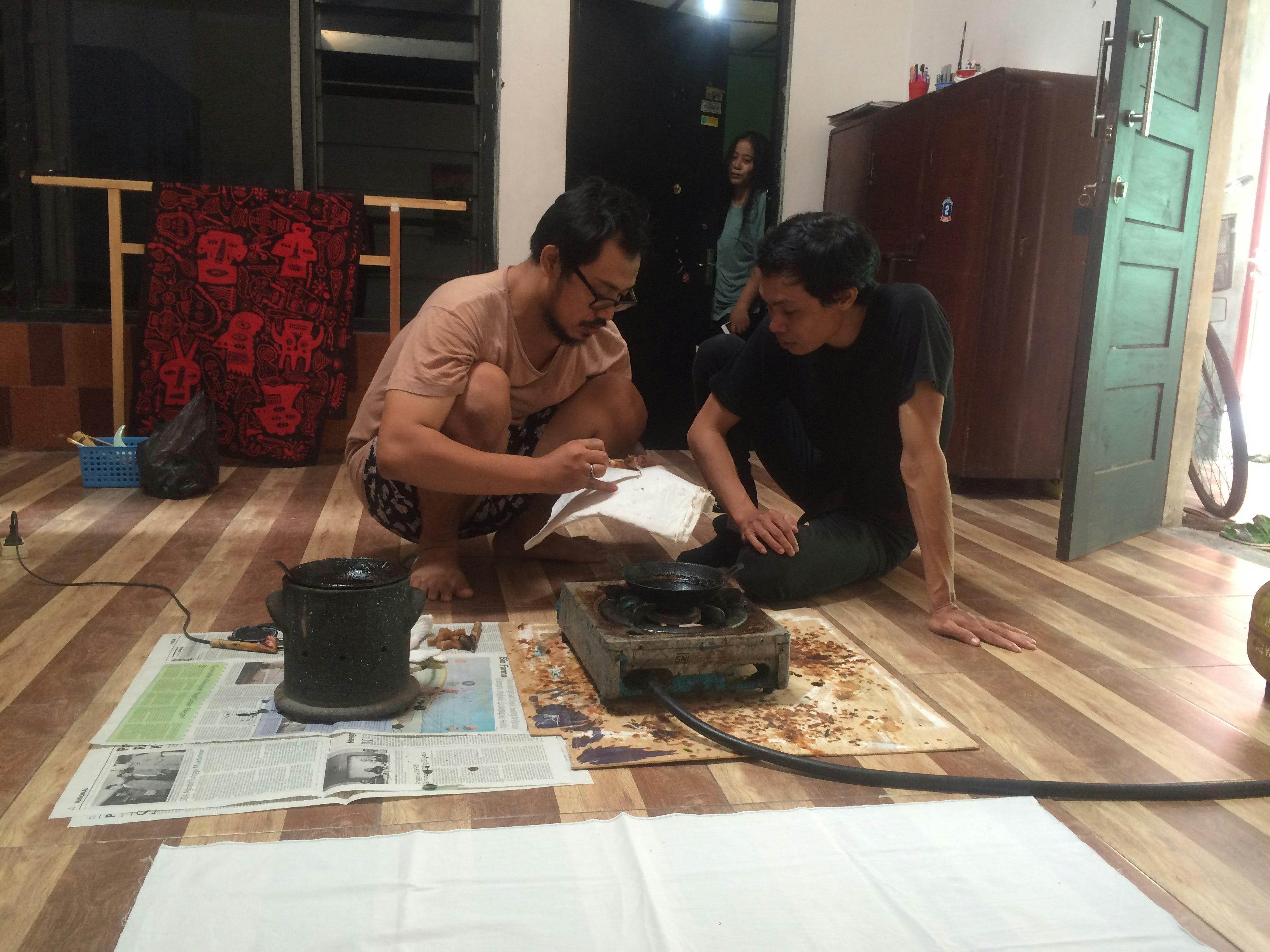 Two male-presenting figures squat on the ground over a gas stove.