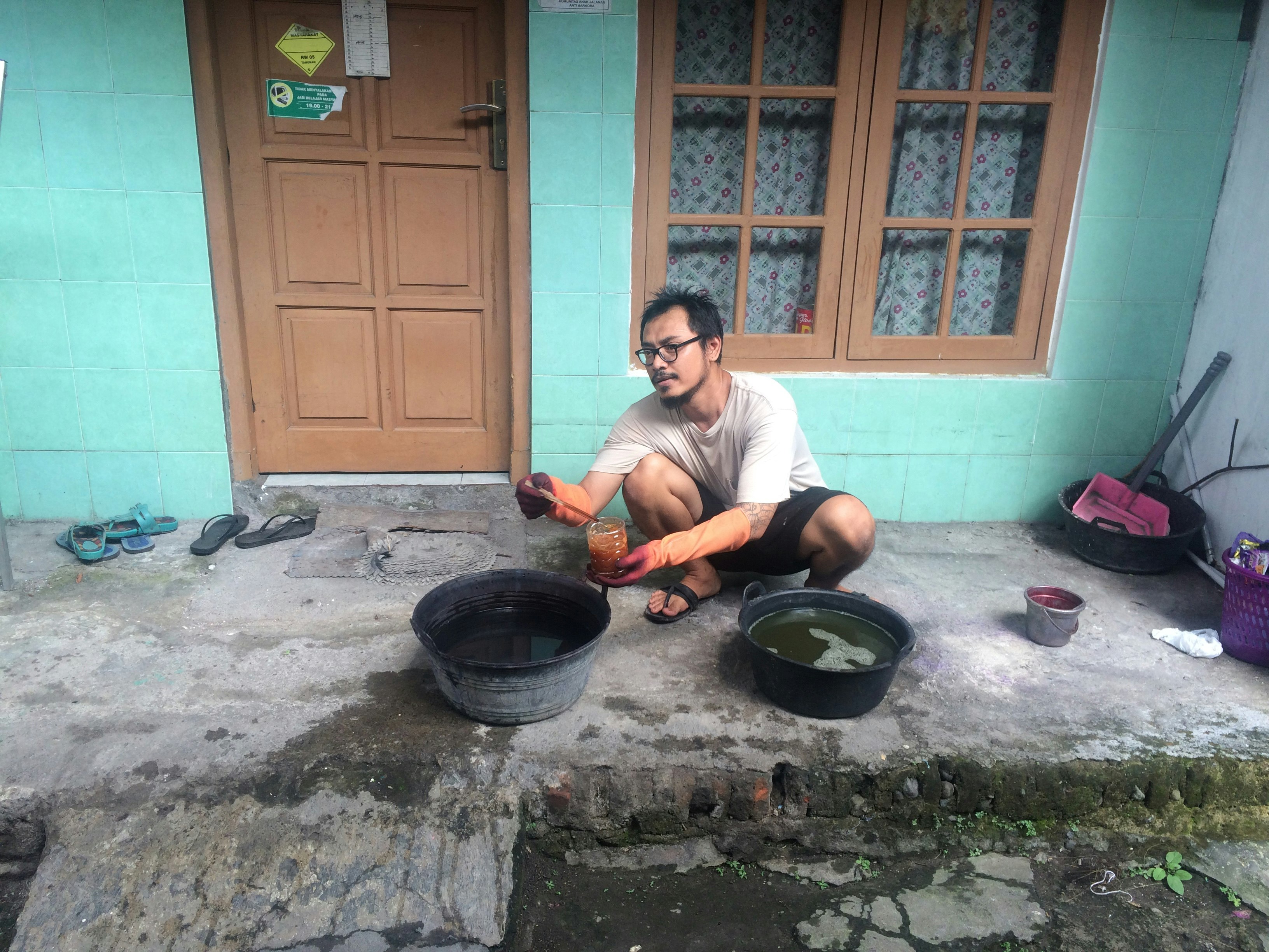 Arwin, a male-presenting Indonesian artist, squats over two tubs of liquid in front of a teal green house.