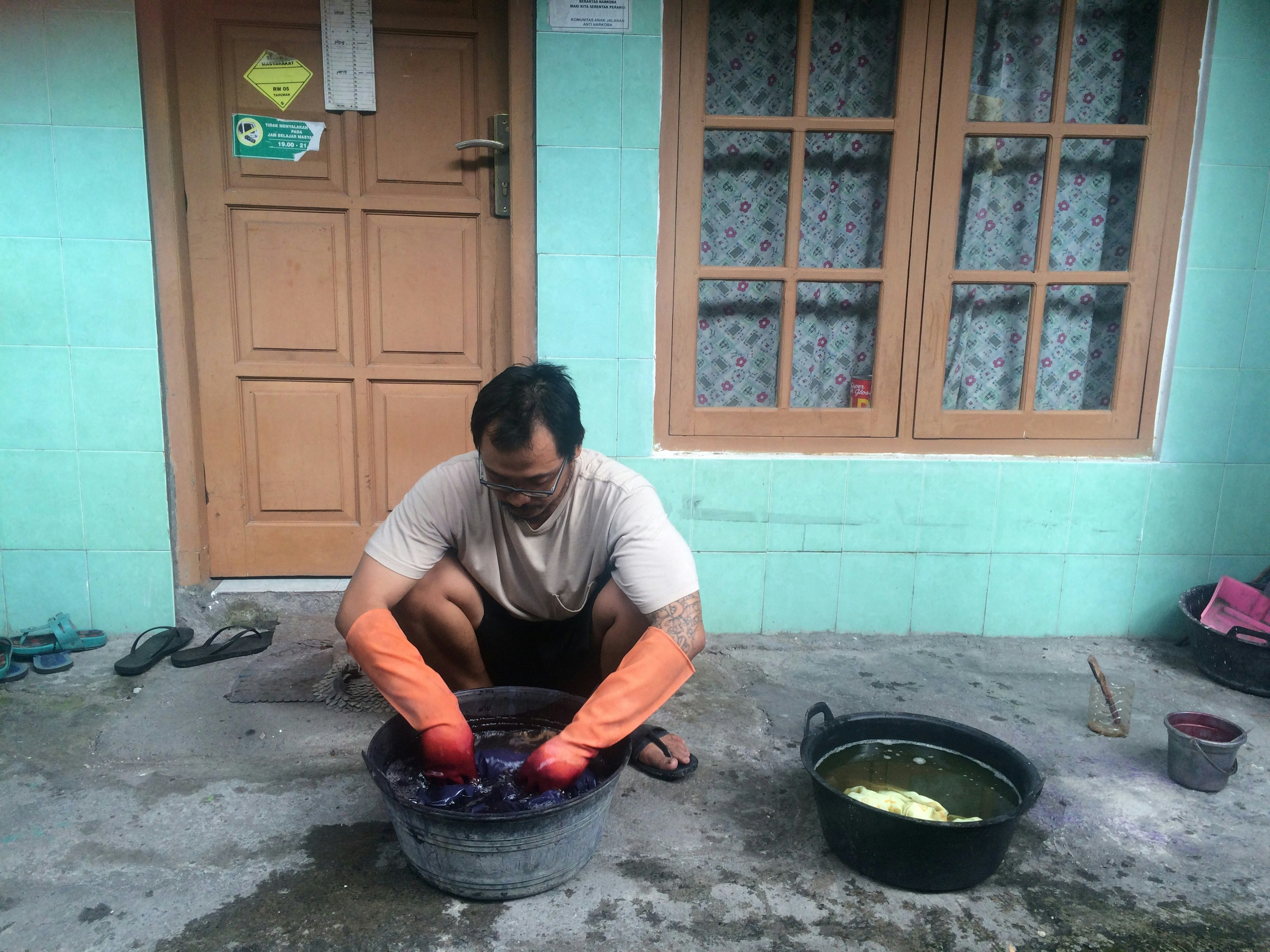 Arwin dips a fabric into a tub of liquid while wearing orange elbow-length dishwashing gloves.