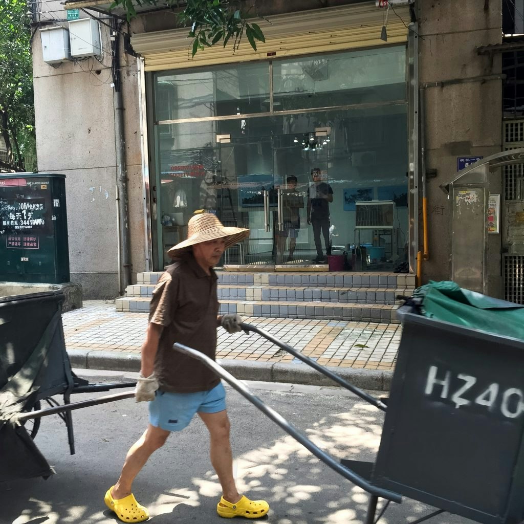 A street hawker wearing a straw hat and yellow Crocs pulls his wares across the front of a glass-front gallery.