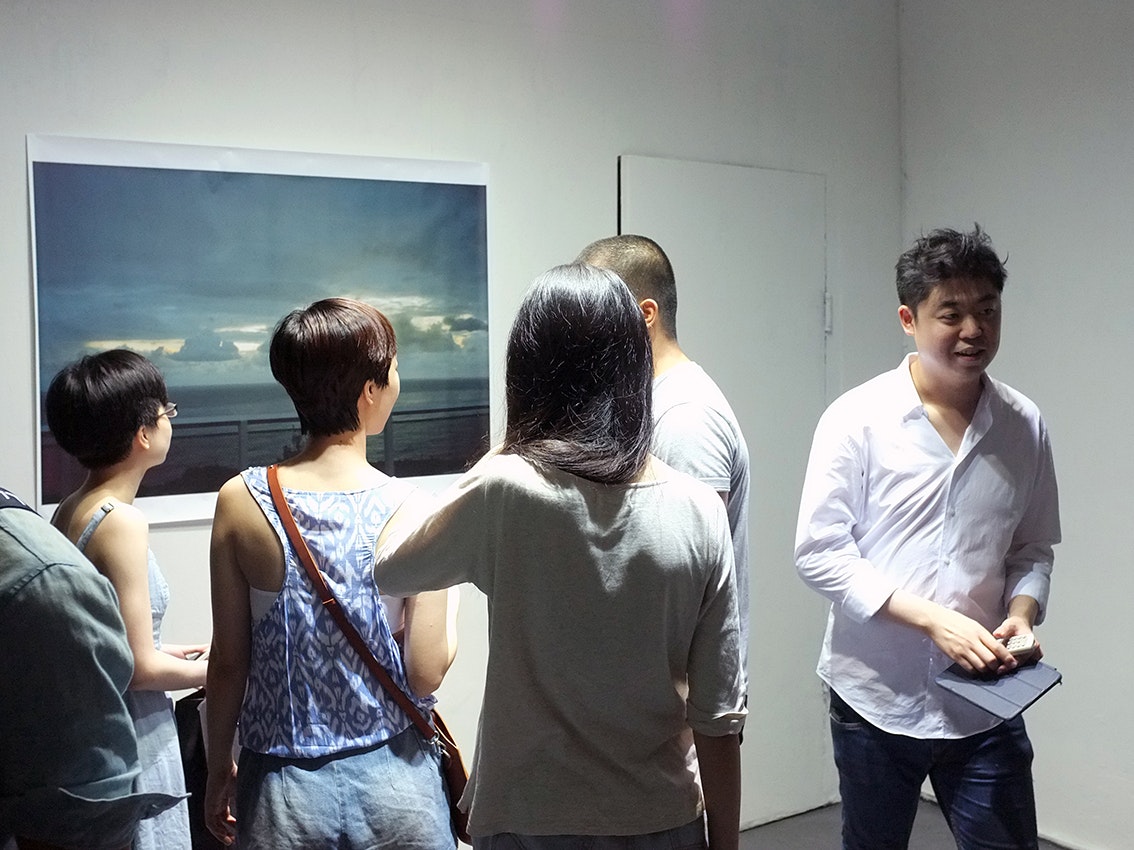 Anthony, an East Asian male-presenting figure wearing a white dress shirt, stands next to a crowd of gallery attendees.
