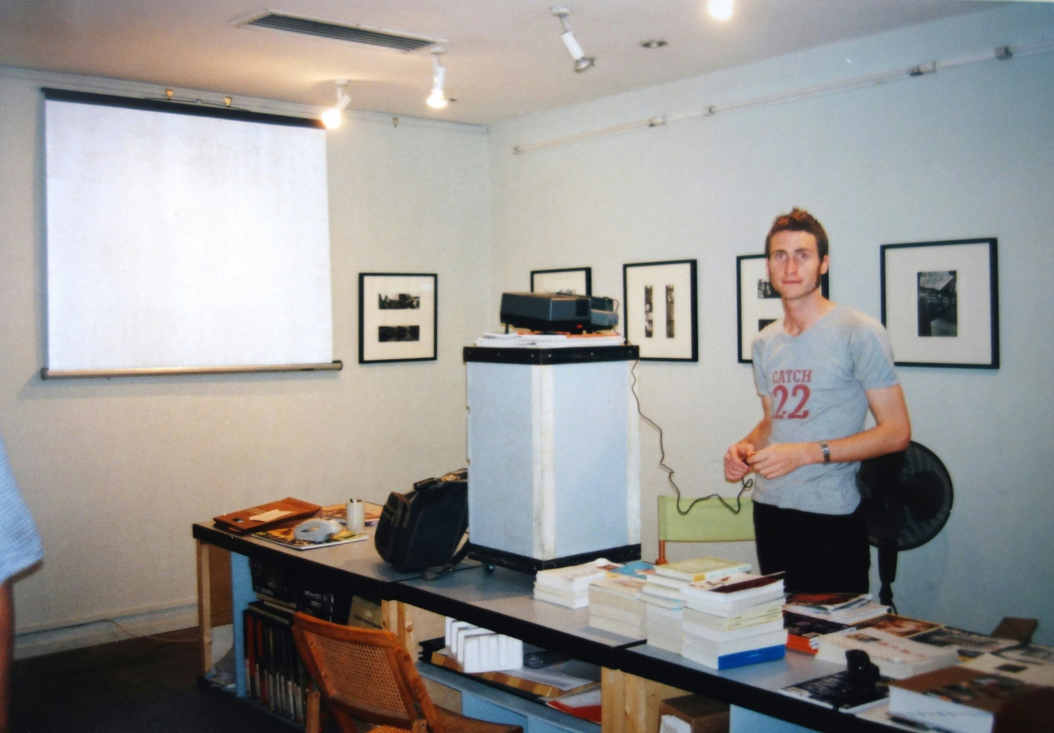 Lucas, a Caucasian male-presenting figure with short brunette hair wearing a grey t-shirt, standing in front of a projector and projector screen.